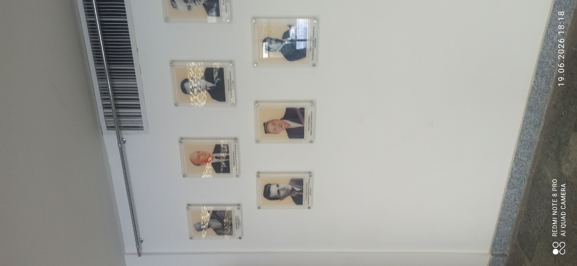 A wall displays several framed portraits of men in formal attire. The portraits are arranged in columns and rows, with some in clear plastic sleeves. Some of the portraits have a shimmering overlay or reflection. A ventilation grate is visible on the left side of the image, with a metal rod running vertically in front of it. The wall is painted white. In the background, on the right side, there is a dark, textured surface with a speckled pattern, and text that reads "19.06.2026 18:18" and "REDMI NOTE 8 PRO AI QUAD CAMERA". The lighting appears to be artificial, as there are reflections on the portraits. The overall scene suggests a formal setting, possibly a hallway or a room dedicated to displaying achievements or historical figures.