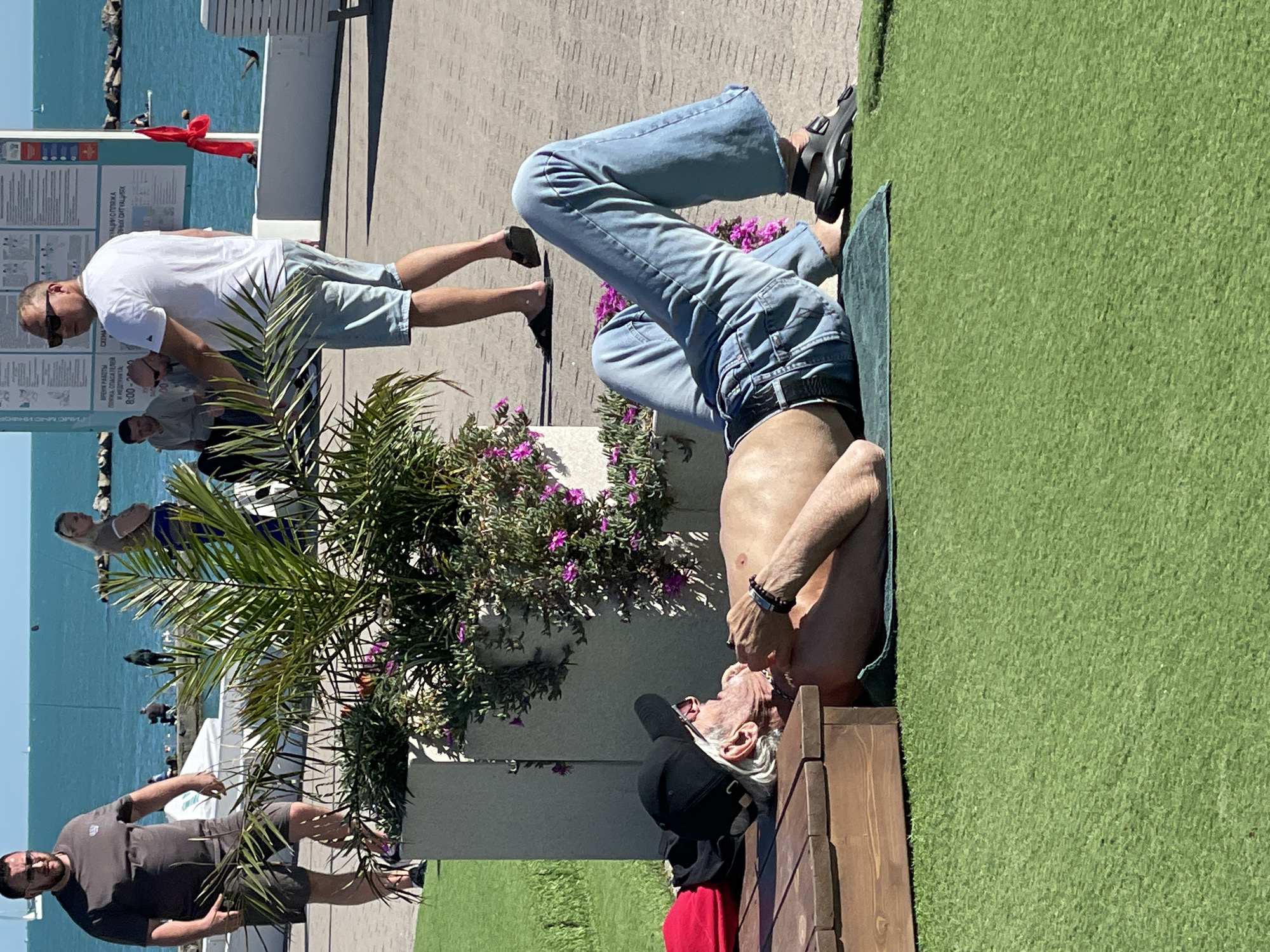 This media file depicts an outdoor leisure scene on a bright, sunny day at a waterfront promenade, consistent with a resort location like Sochi, Russia. The foreground features artificial green turf, where a shirtless man is relaxing on a blue towel beside a wooden bench. He wears a black cap and appears to be lying on his back, looking upwards. He is wearing blue jeans.
To the left, another man in a white t-shirt, light blue shorts, and sunglasses stands on a paved area, looking down. A vibrant palm tree with green fronds and purple flowering shrubs are centrally placed, separating parts of the scene. Further back, the clear blue water of the sea or ocean is visible, with several boats in the distance. Additional people are scattered throughout the background, including a man in a dark shirt and shorts in the bottom left.
Informational signs written in Russian are present near the waterfront. Visible text includes "ВРЕМЯ РАБОТЫ ПЛЯЖА, СПАСАТЕЛЕЙ И МЕДПУНКТАС" (Operating hours of the beach, lifeguards, and first-aid post) followed by "8:00-20:00". Other fragments read "Тимс МНС ИНО" (likely "МЧС ИНФО" for Ministry of Emergency Situations Info) and parts of phrases related to "эвакуации с пляжа" (evacuation from the beach) and "чрезвычайных ситуациях" (emergency situations). The bright sunlight and clear sky indicate excellent weather, contributing to a relaxed, recreational atmosphere.
