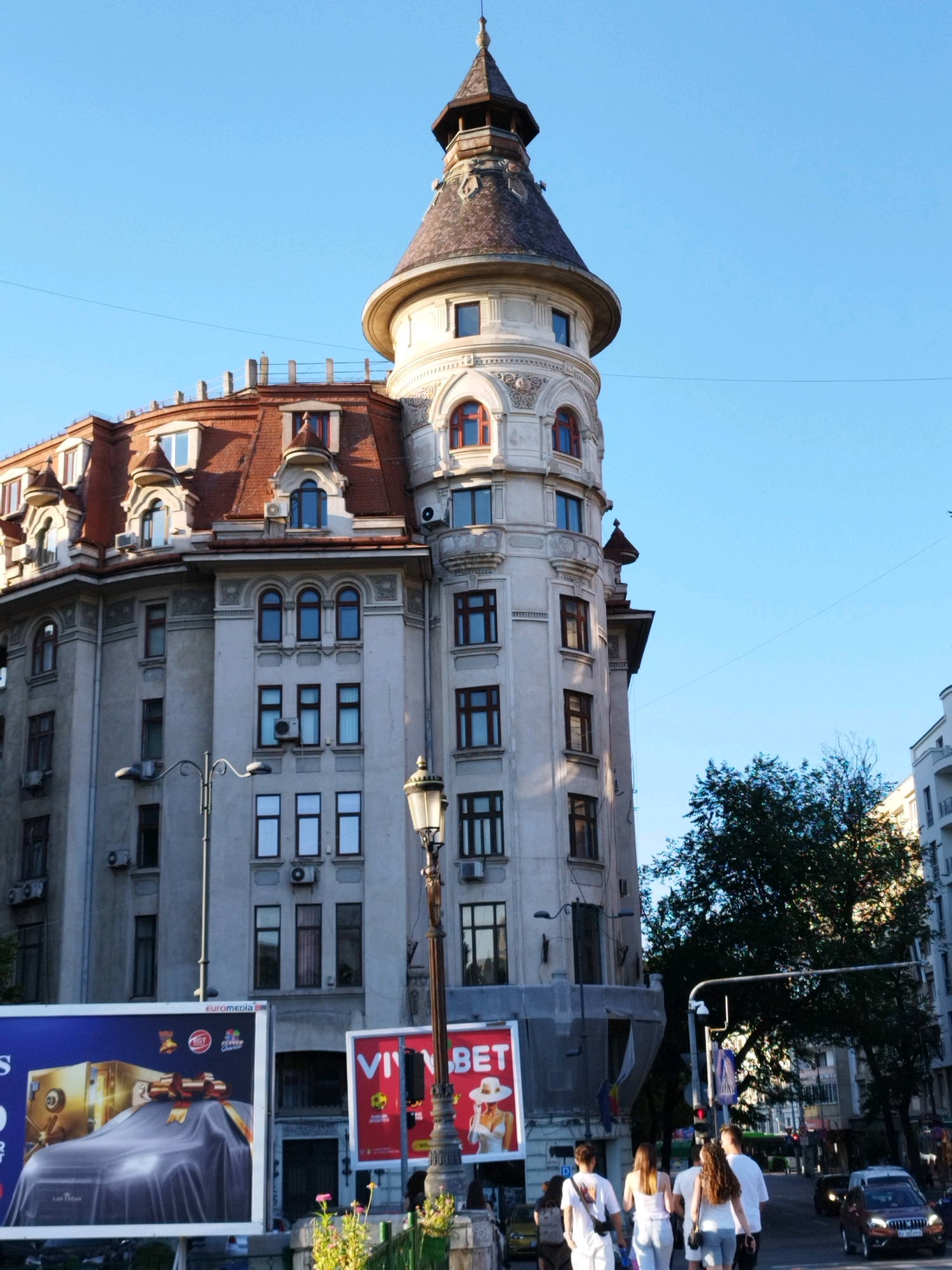 The image captures a sunlit urban street scene in Bucharest, Romania, dominated by a distinctive, ornate building with a conical tower. The building's facade is a pale gray, adorned with various architectural details including arched windows, decorative moldings, and a prominent red-tiled roof. Several air conditioning units are visible on the building's exterior. In the foreground, two large billboards are displayed. The left billboard is blue and features an image of a car covered by a draped cloth, next to a golden safe. The right billboard is red and white, displaying a woman in a white hat.
A lamppost with multiple light fixtures stands to the left of the building, and another ornate lamppost is situated to the right, closer to the foreground. A group of four young adults, two males and two females, are walking away from the camera on the sidewalk. They are casually dressed. The sky above is a clear, bright blue. Trees and other buildings are visible in the background, suggesting a densely populated urban environment. The overall impression is of a bustling city on a clear day.