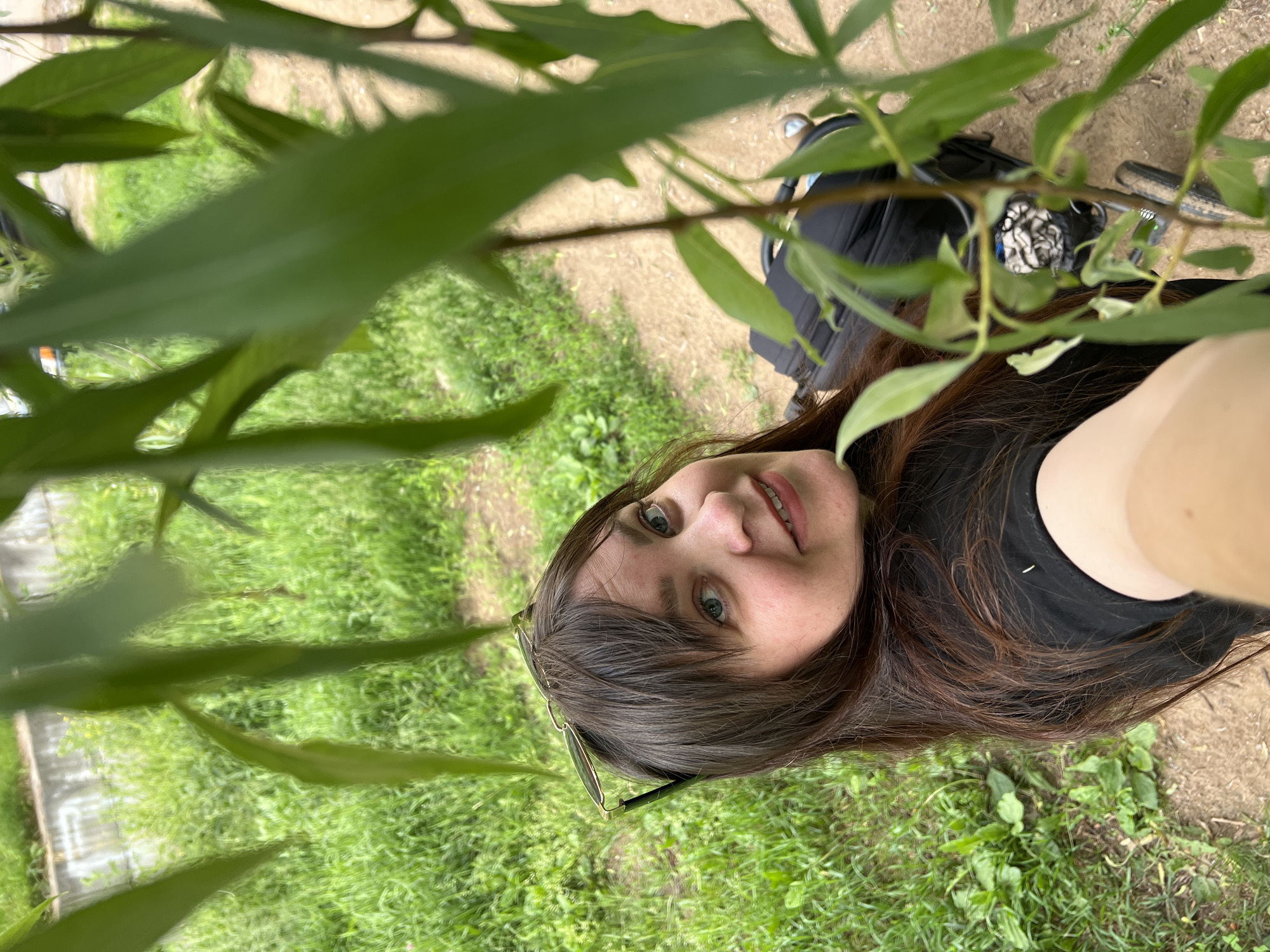 A selfie is taken by a woman with long, dark brown hair and blue eyes, wearing a black shirt and sunglasses perched on her head. She is outdoors, with a background of green grass, a dirt path, and some foliage in the foreground. A black suitcase is visible behind her. The location is the city of Noviy, Russia.