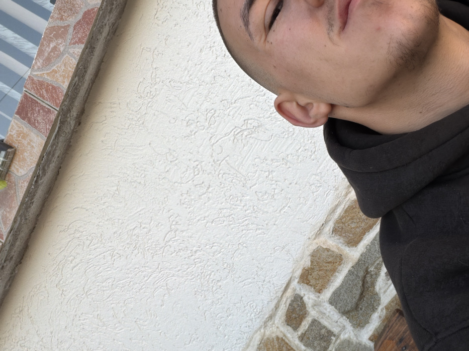 The image displays a close-up, angled view of a male individual in an outdoor setting, potentially in Tarnava, Bulgaria, although no specific visual identifiers confirm this location. The lower right section of the frame features the right side of a male's face, including his right ear, right eye, and portions of his nose and mouth. He exhibits a short, faded haircut on the side of his head and sparse stubble along his jawline and upper lip. He is clad in a dark, likely black, hooded garment. The dominant background element is a light-colored, off-white textured stucco wall. Above this wall, a dark gray concrete or stone ledge runs horizontally, topped by a section of reddish-brown and tan patterned tiles. A narrow strip of a blue and white striped awning or roof element is visible at the very top. A dark wooden surface is partially observable in the bottom left. The scene is illuminated by natural daylight.
