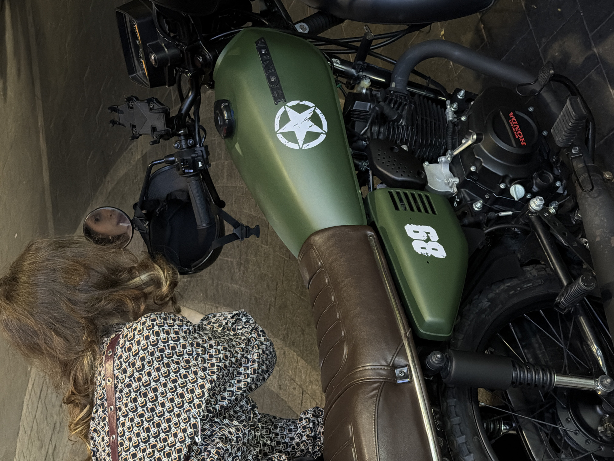 A high-angle, slightly canted view captures a person next to a parked motorcycle. The person, likely a woman, has shoulder-length brown hair and wears a patterned shirt. She appears to be looking at the motorcycle's side mirror. The motorcycle has an olive-green fuel tank marked with a white star and the word "HONDA." A brown leather seat is visible. The motorcycle engine is black. A license plate or number "68" is painted on the side. The location is not identifiable as Đà Nẵng, Vietnam. The surface beneath the motorcycle appears to be stone.