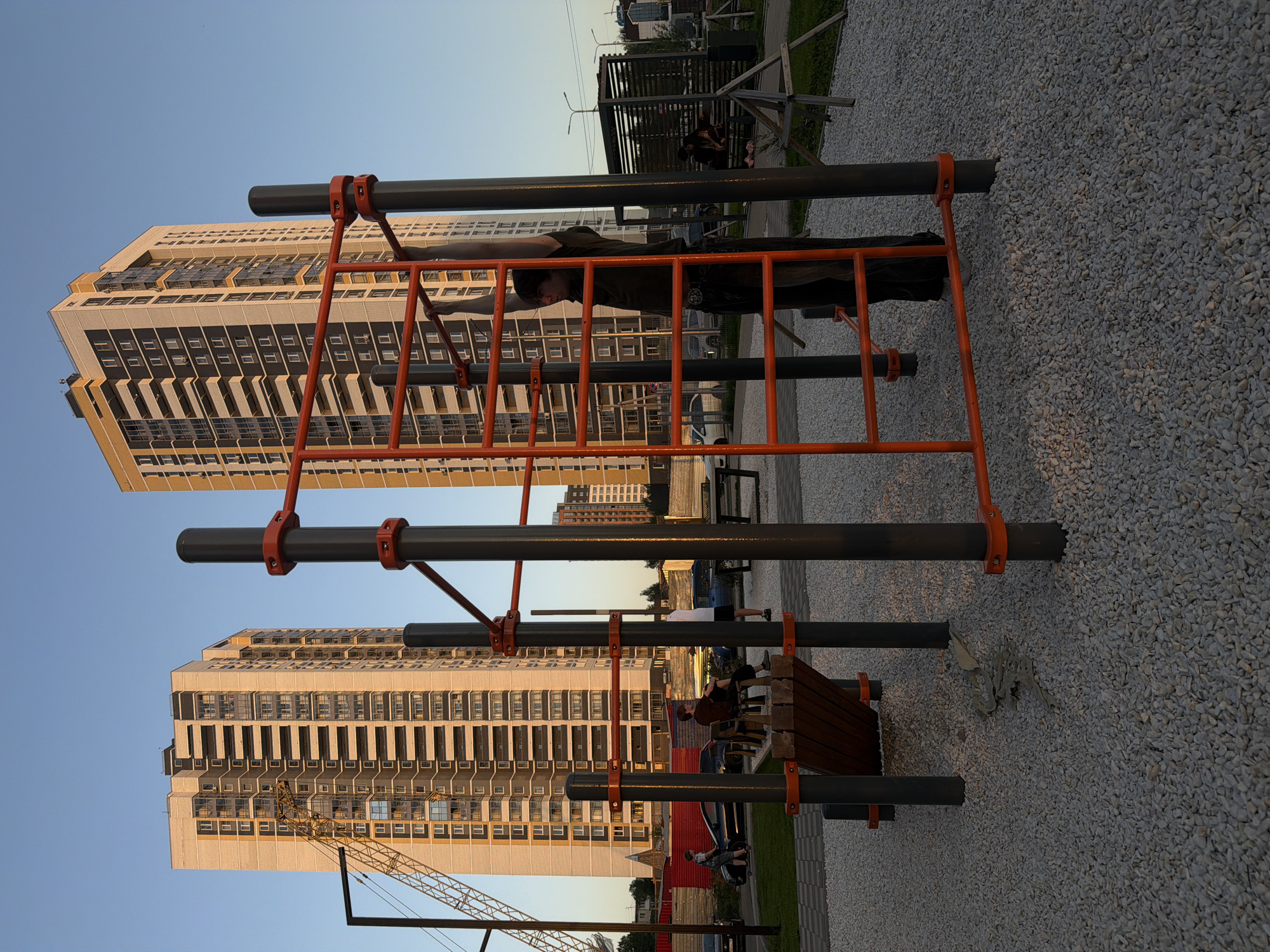 A street workout complex stands in a public park in Izhevsk, Russia. The apparatus is painted in orange and gray. A person is seen hanging from the ladder portion of the complex. In the background, there is a high-rise building with numerous windows. Other people can be seen on benches around the park. The ground is covered in light-colored gravel. In the lower left corner, part of a crane is visible.