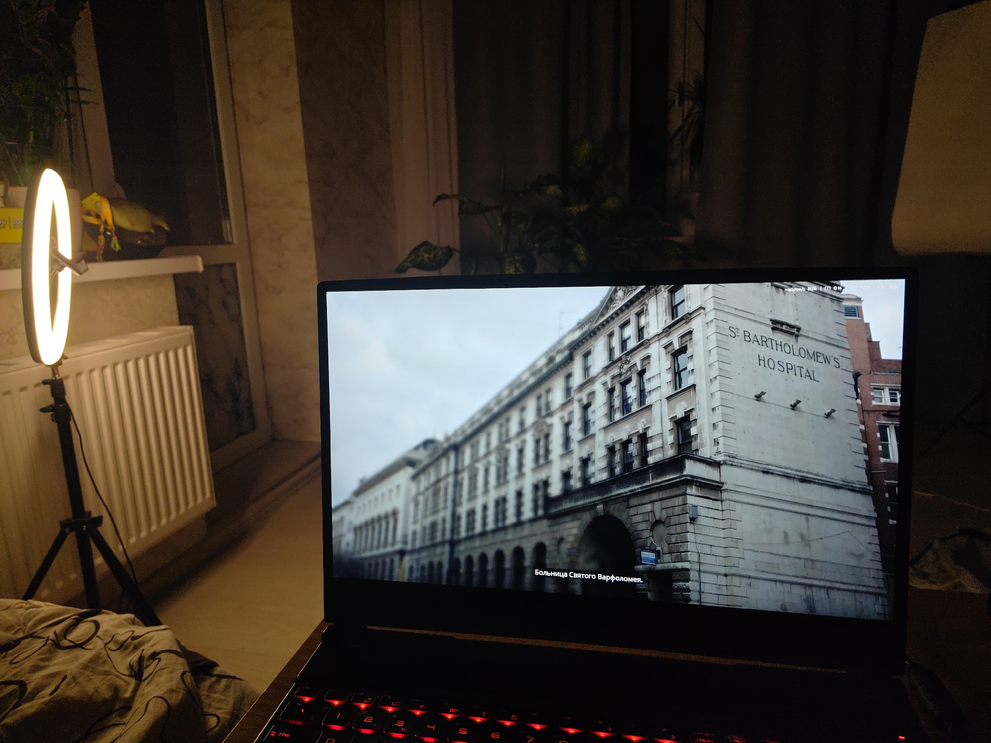 In an indoor setting, a laptop displays a cinematic scene of the St Bartholomew's Hospital in London, England. The hospital building, characterized by its stone facade and arched entrance, is prominently featured. Below the hospital name, Cyrillic text is visible, indicating "Hospital of Saint Bartholomew." To the left of the laptop, a ring light on a tripod illuminates the area. A radiator is visible behind the ring light, and a blurred image of plants and a window can be seen further in the background. The foreground shows a portion of a bed covered with a patterned blanket. The scene suggests an individual is engaged in watching a movie or video on their laptop, possibly in a bedroom or living space. There are no people depicted in the image. The presence of the laptop, ring light, and indoor setting suggests a personal or entertainment-focused activity.