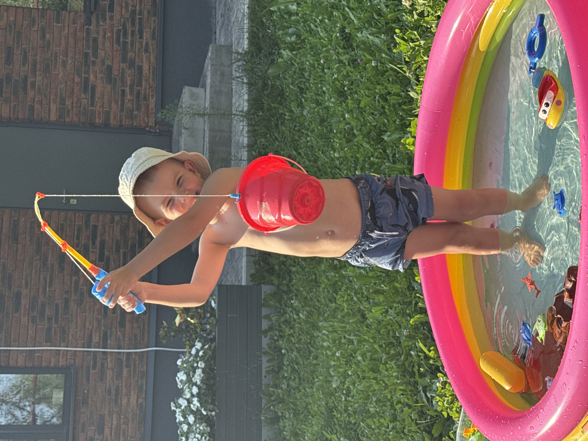 A young boy, wearing a light-colored bucket hat and dark swim trunks, stands in an inflatable kiddie pool filled with water and toys. He is holding a toy fishing rod with a blue reel and yellow and orange pole. A red bucket is suspended from the fishing line. The pool is surrounded by green grass. In the background, there is a brick building with a dark door and a window, and some white flowers in a planter. The scene appears to be a residential backyard. This image does not provide any specific information to determine its location in Chaikovsky, Russia.