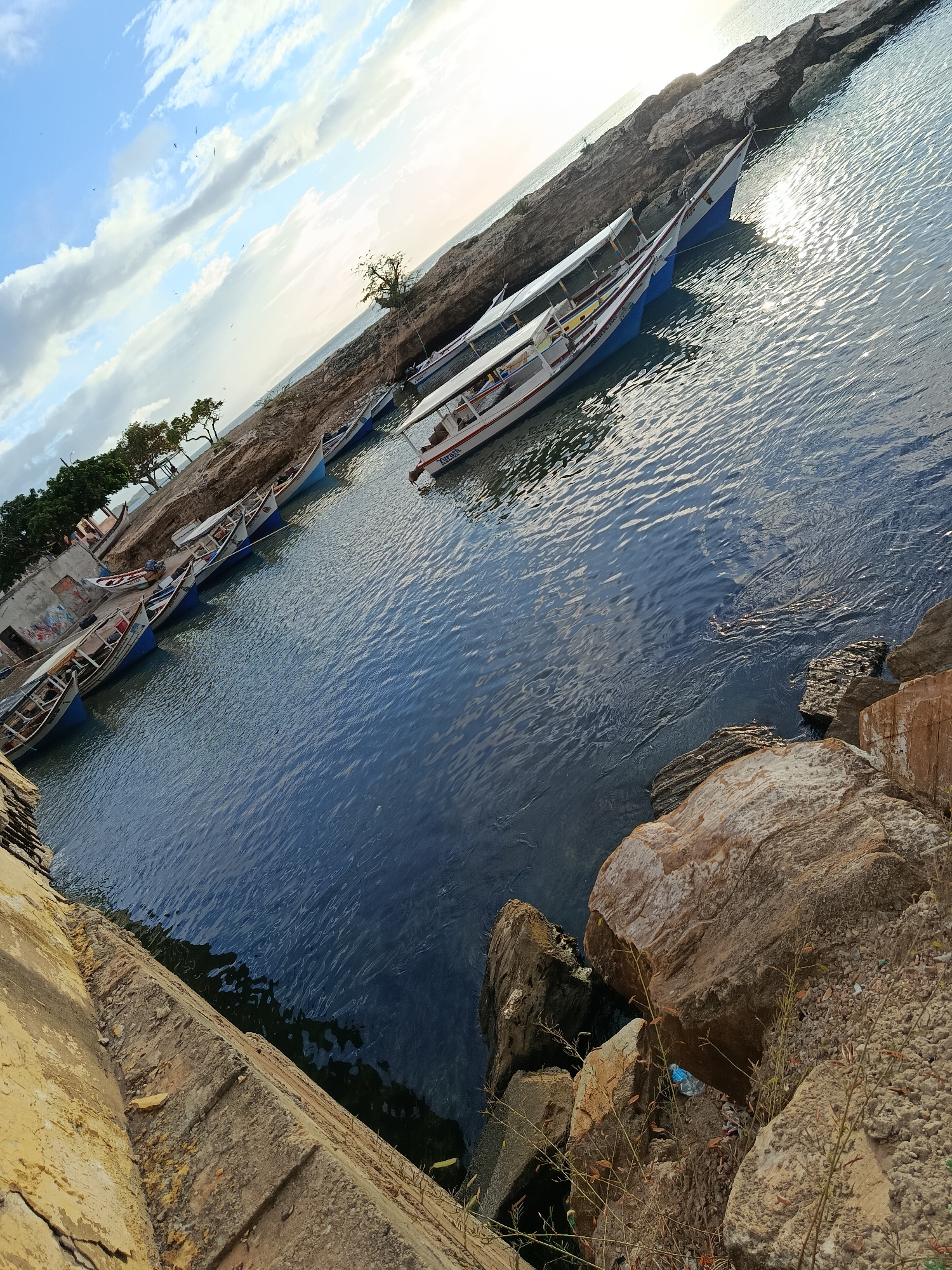 A sun-drenched, angled shot captures the coastline of Juan Griego, Venezuela, featuring several small fishing boats docked near a rocky shore. The water, a deep blue, reflects the bright sky and the sun's rays, creating a shimmering effect. The boats, lined up along the shore, are of varying sizes, with some appearing to be traditional wooden fishing vessels and others more modern tourist boats. One of the tourist boats has "Juan Griego" written on its side. The rocky shore is rugged and uneven, with some vegetation growing out of the crevices. In the background, a few trees and a small, weathered building with graffiti can be seen. The sky is a mix of blue and white clouds, with the sun partially obscured, suggesting it might be late afternoon or early morning. The scene is devoid of any visible human activity, giving a sense of quiet tranquility to the coastal landscape. The foreground is dominated by large rocks and a textured concrete wall, hinting at a man-made structure like a pier or seawall.