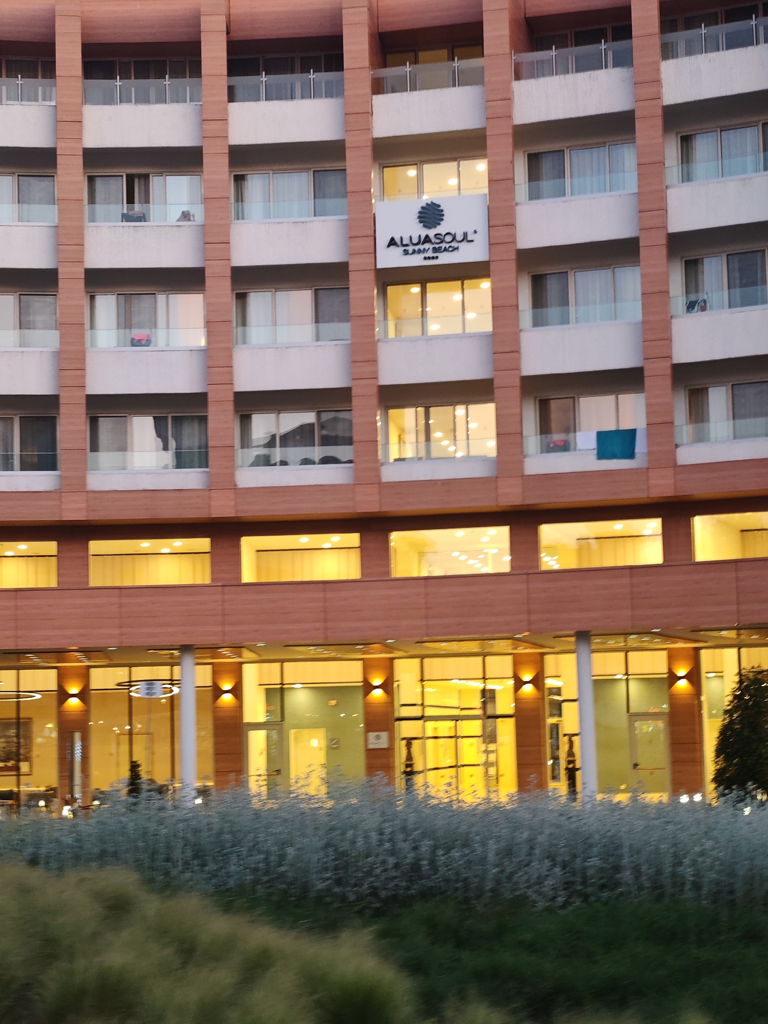 The image shows the exterior of the Aluasoul Sunny Beach Resort in the city of Nesebar, Bulgaria. The building is multi-story with a distinct architectural style, featuring a series of windows and balconies. The hotel's sign is visible. The ground level has a row of windows with illuminated interiors. Bushes and other vegetation are located in front of the building.