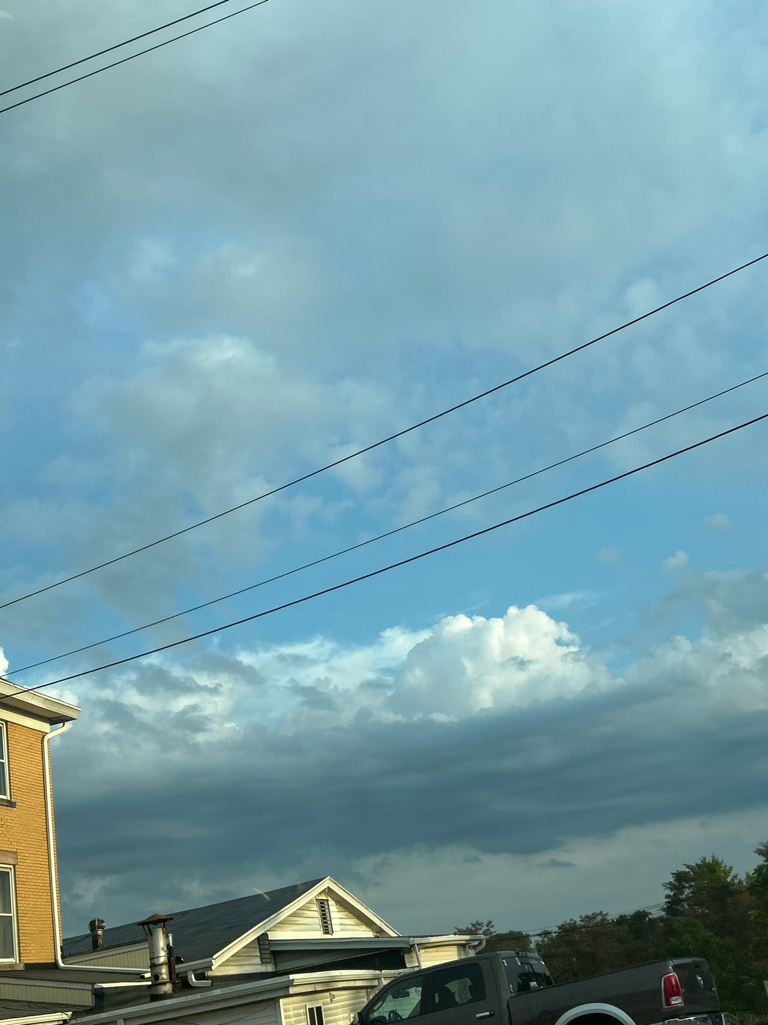 A wide shot captures a suburban scene in Robinson Township, USA, under a dramatic sky. Three parallel power lines stretch across the frame, dividing the canvas into horizontal segments. The upper portion of the image is dominated by a gradient of light to medium blue sky, scattered with wispy white clouds. As the eye moves down, the clouds become more dense and darker, transitioning into a deep grey mass that obscures the horizon.
In the foreground, the tops of several houses are visible. On the left, a portion of a tan brick building with white trim and windows peeks into the frame. To its right, a white-sided house with a dark grey roof features a prominent gable vent and a red-capped chimney. Another white structure with a sloping roof is partially visible beneath it. A dark grey pickup truck, facing right, is parked in front of these buildings, its rear bumper and tailgate evident. The tail lights and part of the bed are visible. The scene is bordered by the tops of trees on the far right, suggesting a wooded or park-like area beyond the residential structures. The overall impression is one of an overcast day, possibly with impending rain, offering a contrast between the bright sky above and the dark, heavy clouds.