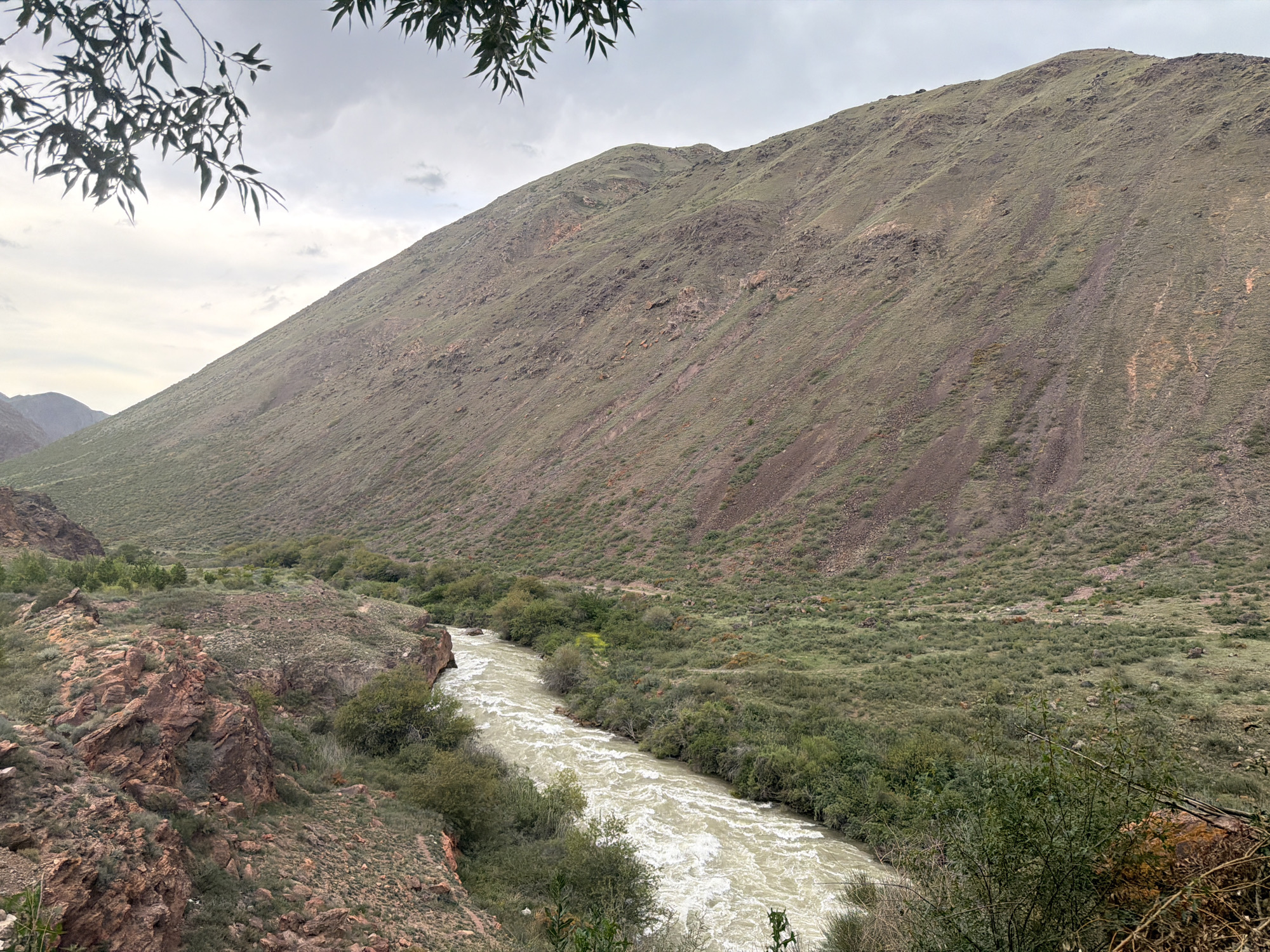 A wide, muddy river flows through a valley with steep, grassy slopes on either side. The river is fast-moving, with whitewater rapids visible in the foreground. The slopes are covered in a mix of grass, shrubs, and rocks. In the distance, the valley narrows, and the slopes become more rugged. The sky is overcast, with clouds obscuring the sun. The overall impression is one of a wild and remote landscape. The image is taken from a high vantage point, looking down into the valley. The time of day appears to be late afternoon or early evening, as the light is soft and diffused. There is no visible text in the image.
