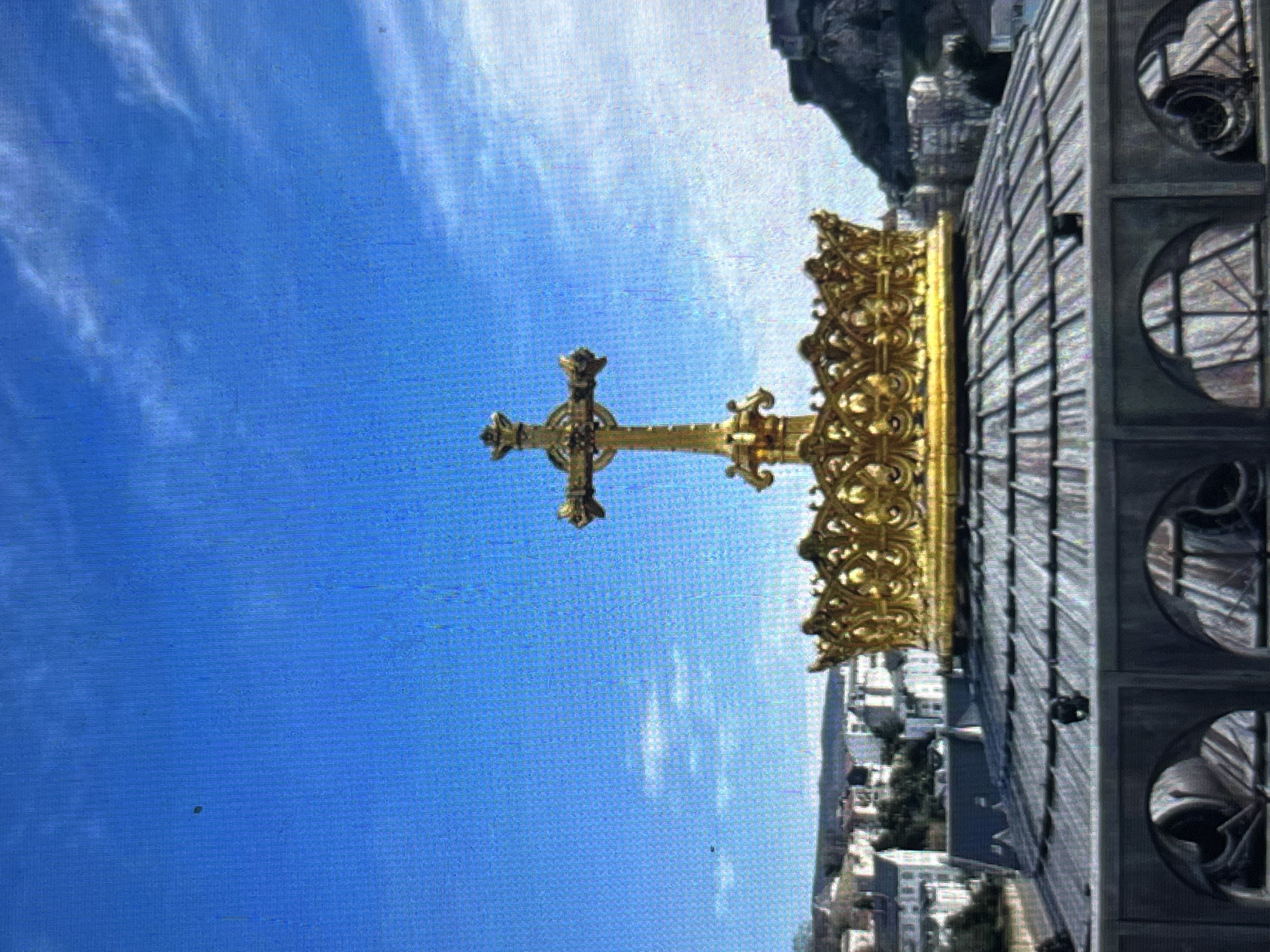 The image depicts a golden ornate cross and crown atop a building in Lourdes, France, set against a blue sky with scattered clouds. Adjacent to the cross are architectural elements, including metal grating and arched windows. In the background, a cityscape is visible with buildings and green spaces. A mountain range appears in the upper right corner.