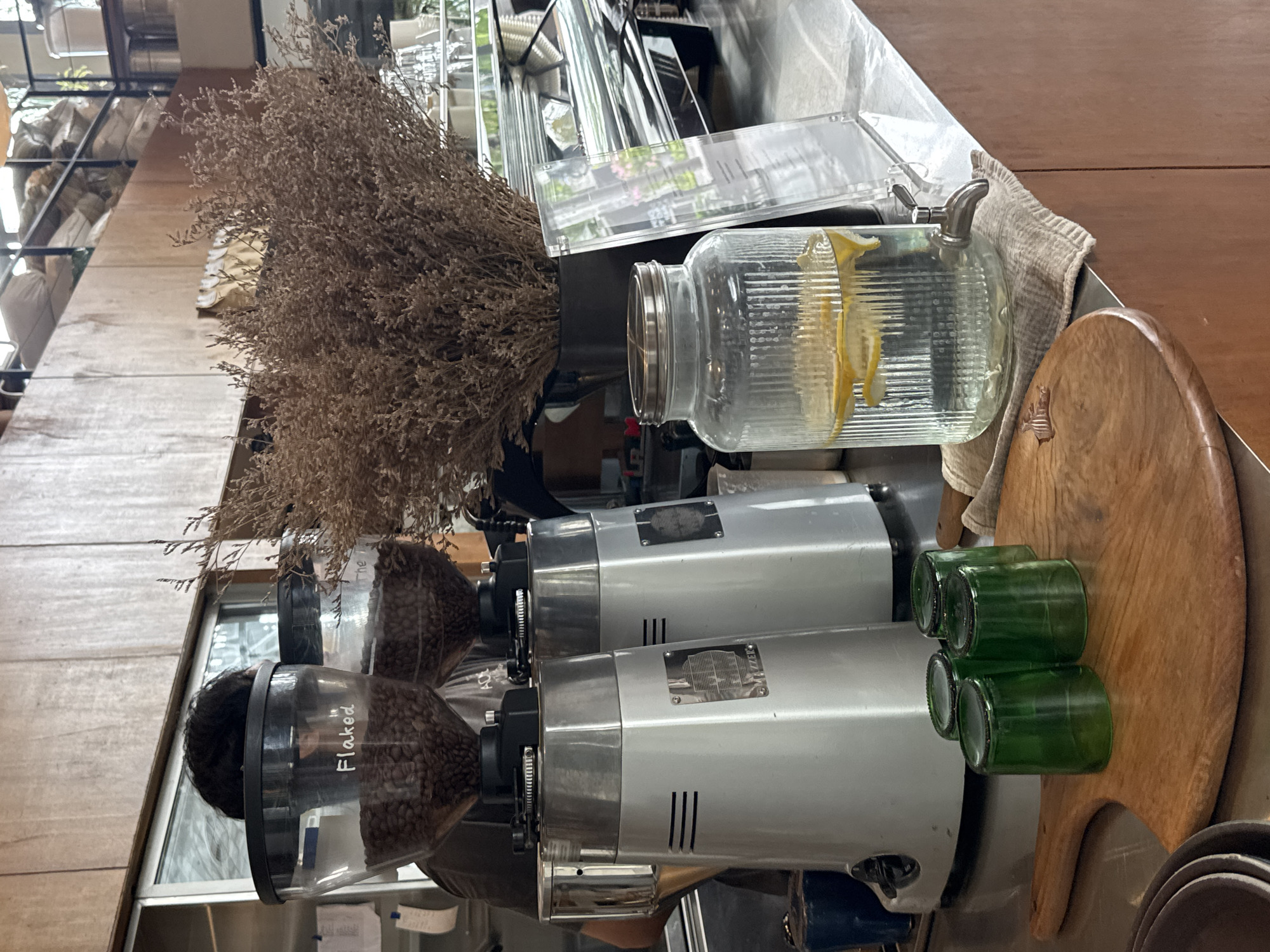 The image shows a countertop scene, possibly in a cafe, in North Kuta, Indonesia. Two commercial coffee grinders are positioned prominently, with clear containers filled with coffee beans labeled "Flaked" and "The". A large glass jar filled with water and lemon slices is visible. Adjacent to the grinders, there is a wooden serving board with four small, green glass containers. Dried floral decoration adds to the ambiance. The background features a shelving unit with various items, partially visible through a glass panel.