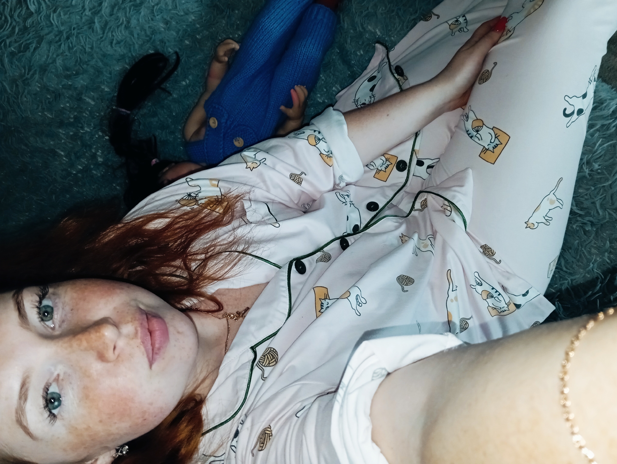 The image shows a woman lying on a fluffy, blue surface. She is wearing pink pajamas with a pattern of cats and yarn balls. A blue doll with a black ponytail also lies on the surface next to her. The woman has red hair, freckles, and blue eyes. She is wearing a gold necklace and bracelet. The location of the image in Yurgamysh, Russia is not identifiable based on the visual information provided.