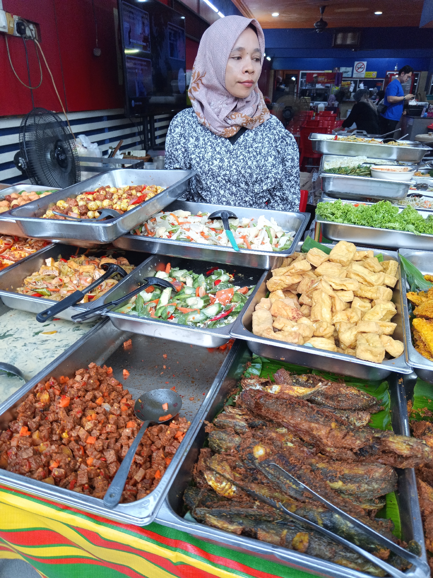 This image depicts a food stall in Kuala Lumpur, Malaysia, likely an outdoor or semi-enclosed market setting. A woman wearing a hijab stands behind a display of various prepared dishes. The food is arranged in stainless steel trays, showcasing a variety of options, including fried fish, fried tofu, stir-fried vegetables, and a dish with cubed meat and vegetables. Serving utensils like ladles and tongs are present in several trays. In the background, a television screen is visible, along with other patrons and shop infrastructure, suggesting a bustling commercial area. The overall atmosphere suggests a local eatery offering a diverse range of Malaysian cuisine.