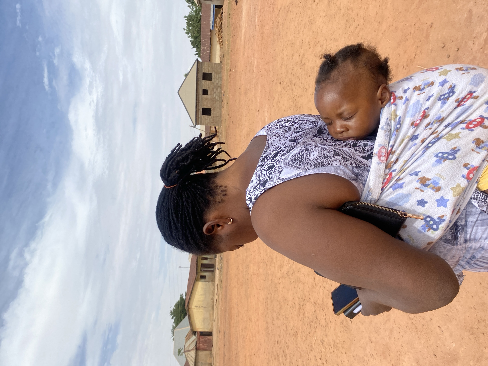In an outdoor setting in Rido, Nigeria, a Black woman with braided hair tied back with a red tie is carrying a sleeping baby secured in a printed white and blue sling. The woman wears a white and black patterned top and has a small gold hoop earring. She is holding a black clutch and a mobile phone in her arms. The ground is reddish-brown soil. In the background, there are simple, one-story buildings, one under construction with exposed brick. The sky is overcast with scattered white clouds.