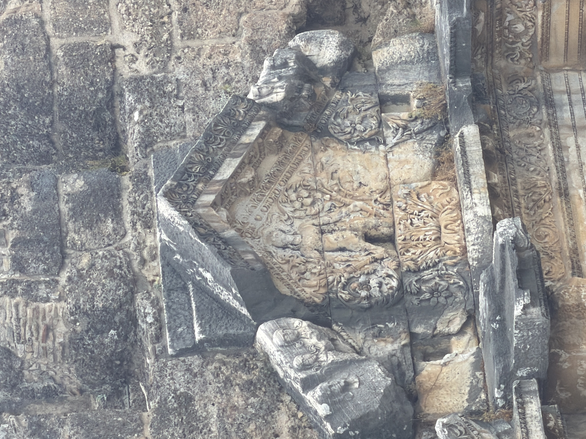 The image displays a close-up view of ancient stone carvings, likely part of a ruin in Serik, Turkey. The stonework is weathered and shows intricate details of relief sculptures. Some carvings appear to depict human faces and floral motifs, while others are more abstract or damaged. The stones are varied in color, ranging from gray to beige, and exhibit signs of erosion. The overall impression is one of historical preservation and decay, characteristic of archaeological sites. The backdrop is a rough stone wall, also showing significant weathering and age.
