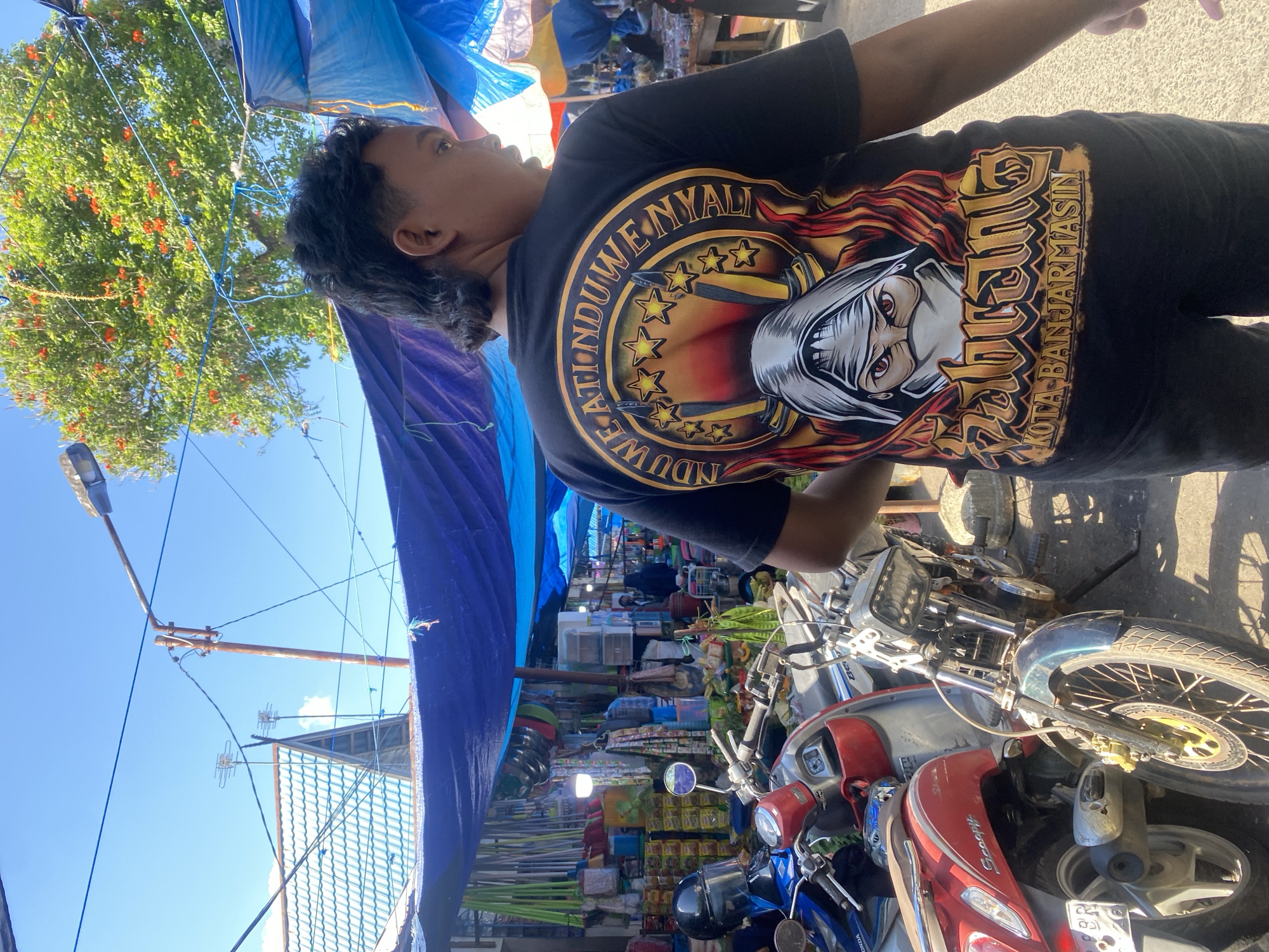 The image captures a street market scene in Desa Tunggul Irang, Indonesia. A person wearing a black t-shirt with a graphic design is the central subject, viewed from behind and slightly to the side. The t-shirt features a circular emblem with stars, swords, and a hooded figure, along with text that reads "NDUWE ATI ND UWE NYALI" and "KOTA BANJARMSIN."
The background reveals a bustling market with blue tarps providing shade over stalls filled with various goods. These include cleaning supplies like brooms and mops, kitchenware such as bowls and colanders, and packaged food items. Several motorcycles are parked amidst the stalls, suggesting a common mode of transportation for vendors and shoppers. One red scooter with the "Scoopy" logo is prominently visible on the left. The environment is characterized by bright sunlight, casting shadows and illuminating the scene. The overall impression is one of a lively, open-air marketplace.