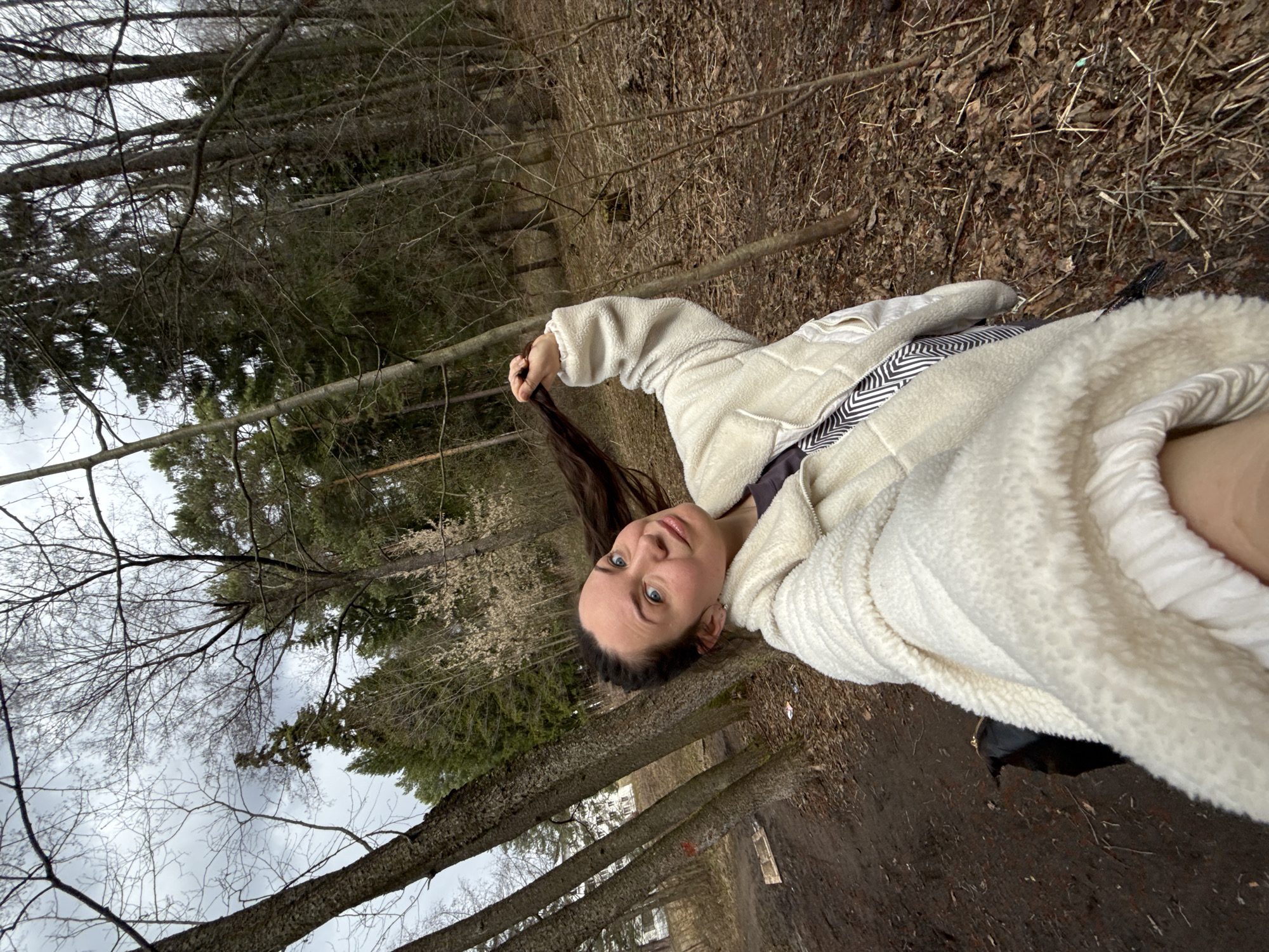A young woman is captured in a close-up, waist-up shot, outdoors in a forest or wooded area in Chaikovsky, Russia. She has long dark hair and blue eyes, and is looking directly at the camera with a gentle expression, holding a section of her hair with one hand. She is wearing a thick, cream-colored, fluffy jacket, possibly fleece or sherpa, over a darker top with a black and white zigzag patterned strap, likely from a bag.
The setting is a natural environment, characterized by numerous trees. The background features both bare deciduous trees and some green conifers, suggesting late autumn, winter, or early spring conditions. The ground is covered with a layer of dry leaves, twigs, and dark soil, indicative of a forest floor. The sky is overcast, providing soft, diffused lighting with no harsh shadows visible. The overall mood is calm and natural. There is no visible text in the image.