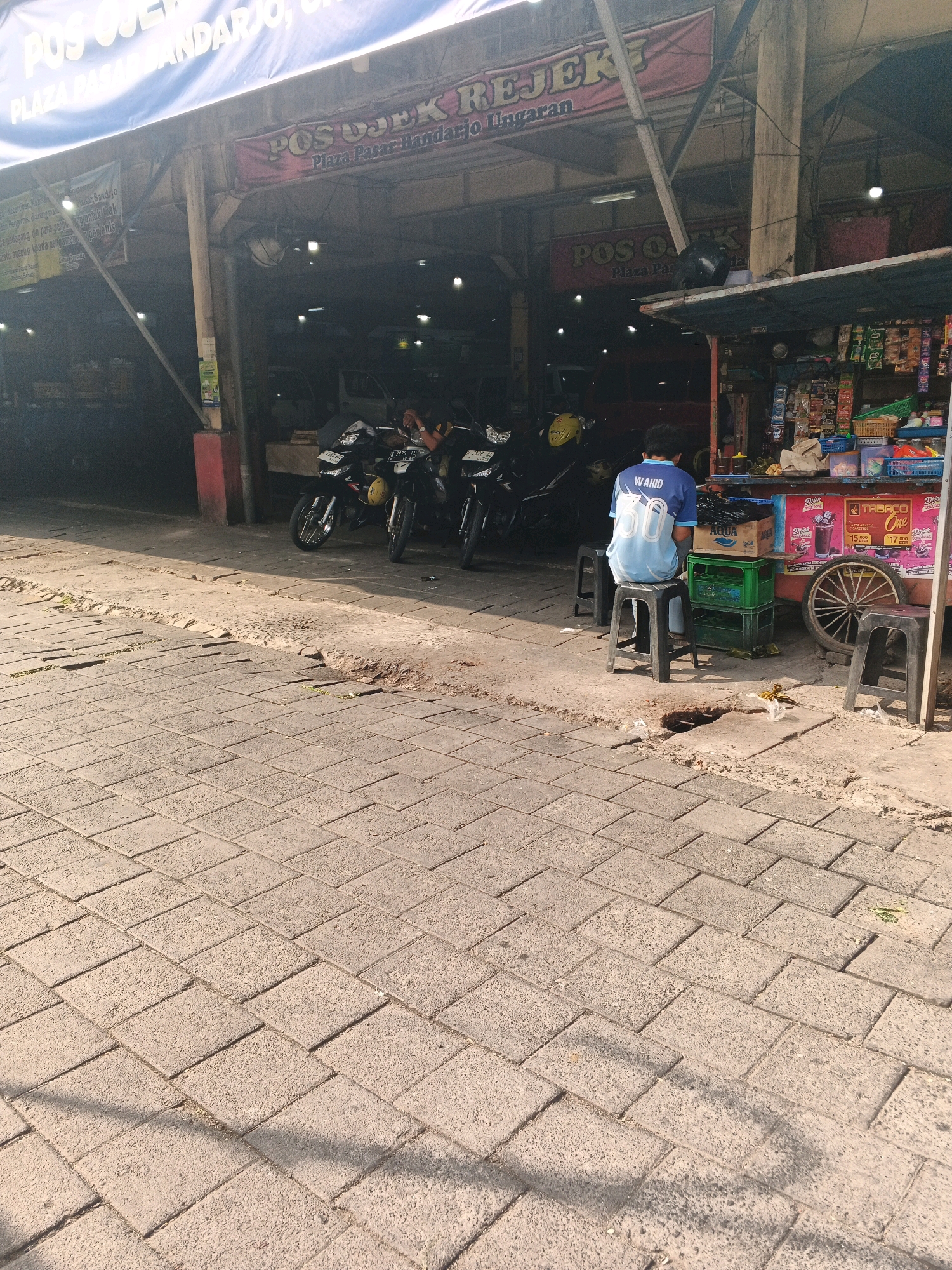 The image captures a street scene in Ungaran, Indonesia, likely near a transportation hub or market given the presence of parked motorcycles. In the foreground, a paved walkway with a brick pattern is visible, with some areas showing wear and tear and patches of dirt. Sunlight casts shadows across the walkway.
In the midground, several motorcycles are parked. Behind them, a covered stall or shop is set up. A young man, wearing a blue and light blue t-shirt with the number "30" on the back and the word "WAHID" above it, is seated on a black plastic stool. He is facing away from the camera, towards the stall. The stall appears to sell various items, including bottled drinks and packaged goods, with colorful signage. A vintage-looking bicycle wheel is attached to the side of the stall.
Further back, a sign with the text "POS OJEK REJEK" and "Plaza Pasar Bandarjo" is prominently displayed, indicating the location as "Pos Ojek Rejek" at "Plaza Pasar Bandarjo" in Ungaran. The structure of the stall and the presence of overhead lighting suggest it is an active commercial area. The overall environment appears to be an outdoor marketplace or a street with vendor stalls.
