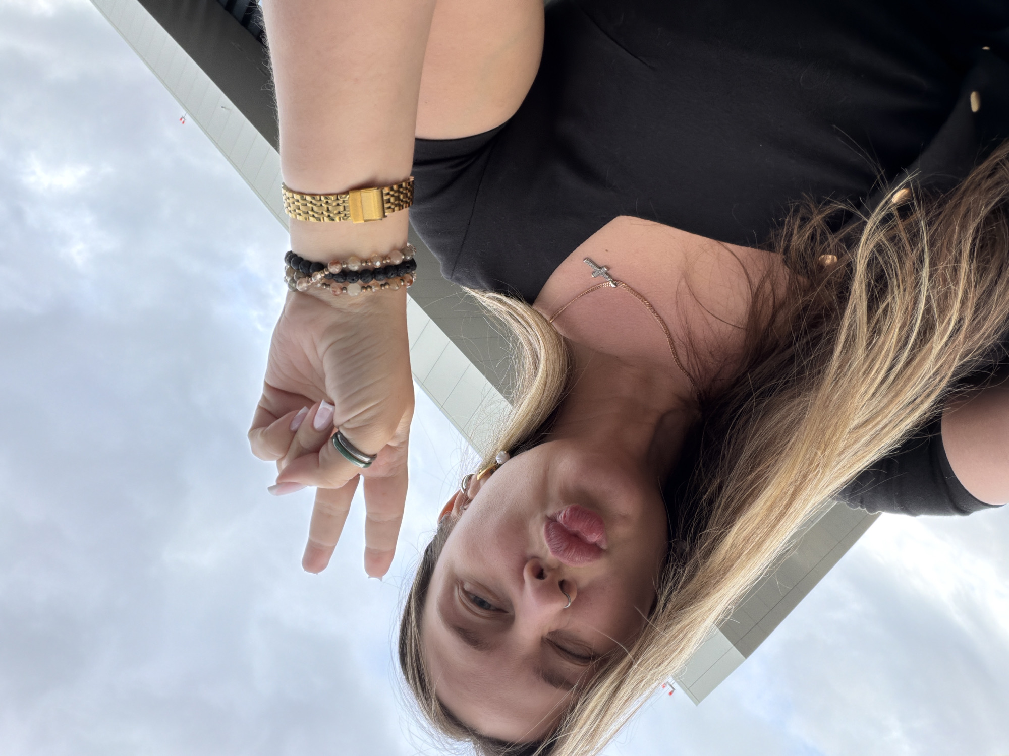 The image captures a close-up, upward-angled selfie of a young adult female located in Staroe Mart'yanovo, Russia. She has light brown hair and wears a black short-sleeved top. Her left eye is squinted, and her lips are pursed in a "kissing" expression. She exhibits a small silver hoop nose ring and multiple earrings on her right ear, including a prominent pearl drop. Her right hand is raised, forming a "peace sign" with extended index and middle fingers, displaying nails with a French manicure. Two thin rings, one silver-toned and one dark green, are worn on her right ring finger. Her right wrist is accessorized with a gold-toned metal link watch and three beaded bracelets of varying colors (dark, clear/amber, and multi-toned). A thin gold-toned necklace with a small, stylized cross pendant rests on her chest. The background consists of an overcast sky with light grey clouds and the upper section of a modern building featuring light grey paneling and a darker grey band, with a small red and white fixture visible on its roofline.