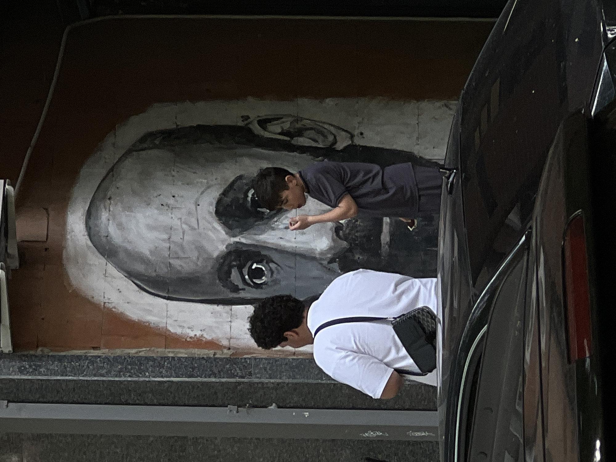 The image shows a large, grayscale mural of a man's face on a wall, likely under a bridge or elevated structure in Yerevan, Armenia. Two young men are positioned in front of the mural. The first man is wearing a dark shirt and appears to be spitting or rinsing something from his mouth towards the mural. The second man, wearing a white shirt and carrying a crossbody bag, is also bending over, possibly observing the action or the mural itself. A dark-colored car occupies the right side of the frame, partially obscuring the view of the scene. The ground appears to be concrete steps or a platform. The lighting suggests an overcast or shaded environment.