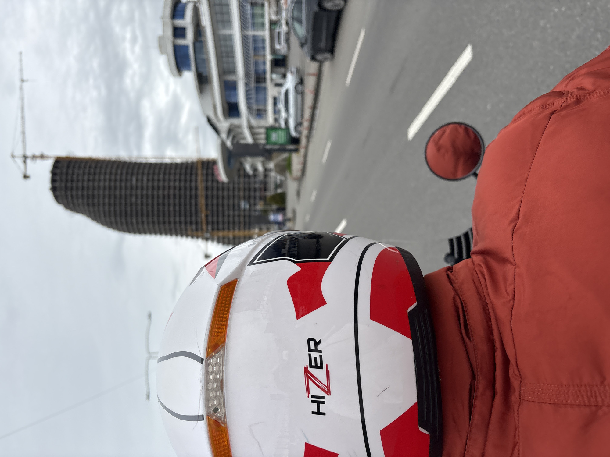 The image depicts a first-person perspective from what appears to be the back of a motorcycle on a city street. The main subject is a white motorcycle helmet with red and black accents and the brand name "HIZER." A rearview mirror is visible on the right side of the frame, along with a portion of an orange jacket worn by the rider. In the background, a city street runs alongside modern buildings, including a partially constructed cylindrical tower. A crane is positioned near the tower. Cars are parked on the side of the road. The sky is overcast. The location is Batumi, Georgia, recognizable by the distinctive architecture of the buildings.