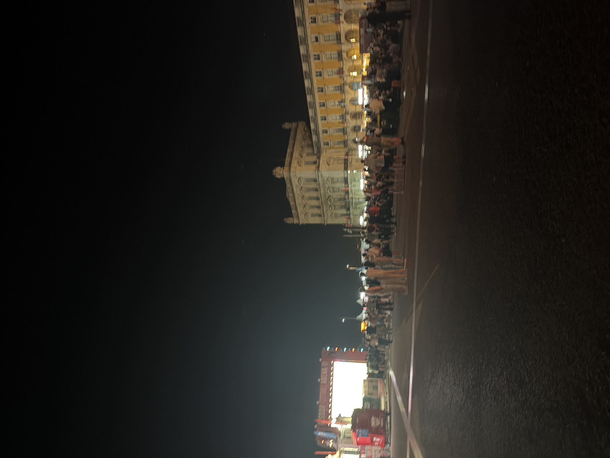 This is a nighttime photograph taken from a low angle in a public square in Lisbon, Portugal. The scene depicts a gathering of people with a large, illuminated screen or stage in the background on the left, labeled "SRIA FESTIVAL ARENA". The screen is rectangular and bright white, with colorful lights bordering it. Adjacent to the screen is a red structure that appears to be a food vendor or kiosk.
To the right of the screen, a crowd of people is visible, standing and mingling. Further to the right, a long, ornate yellow and beige building with multiple windows and arches dominates the background. This building is characteristic of Praça do Comércio, a large public square in Lisbon, Portugal. The square is illuminated by streetlights and the lights from the building itself, creating a warm glow.
More people are gathered in front of the building, some seated on the ground. The overall atmosphere appears to be that of an outdoor event or festival. The ground in the foreground is dark asphalt with white lines, suggesting a paved area like a street or plaza. The sky is dark, indicating it is nighttime, with a few faint lights or stars visible.