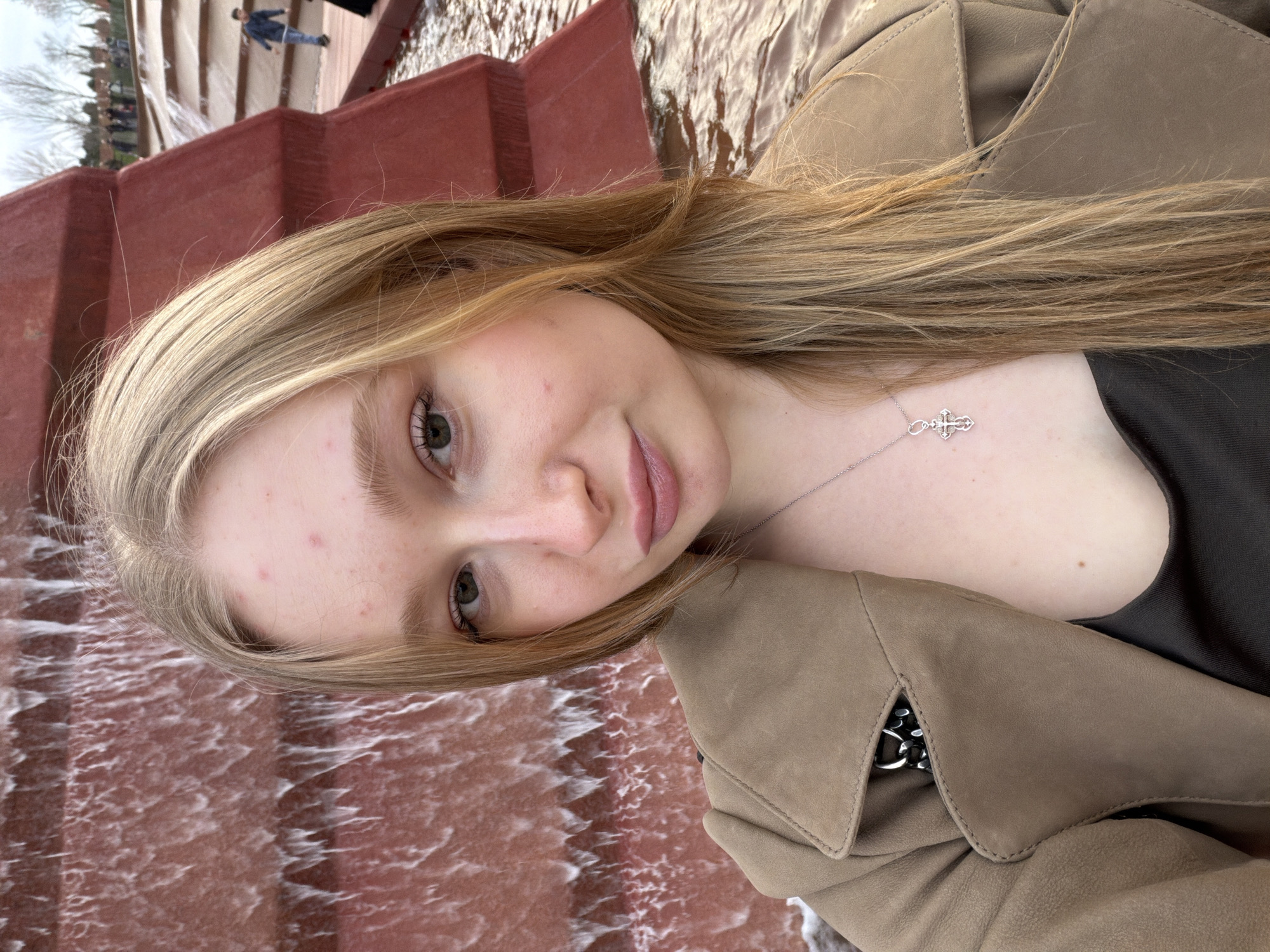 The image shows a young fair-skinned woman with light blonde hair, wearing a tan jacket and a necklace with a cross pendant. She is positioned in front of a cascading red water feature. The background suggests a public space with other people visible in the distance.
Based on the style and color of the water feature, the image is likely taken at the Galitsky Park in Krasnodar, Russia, where this type of water structure is located.