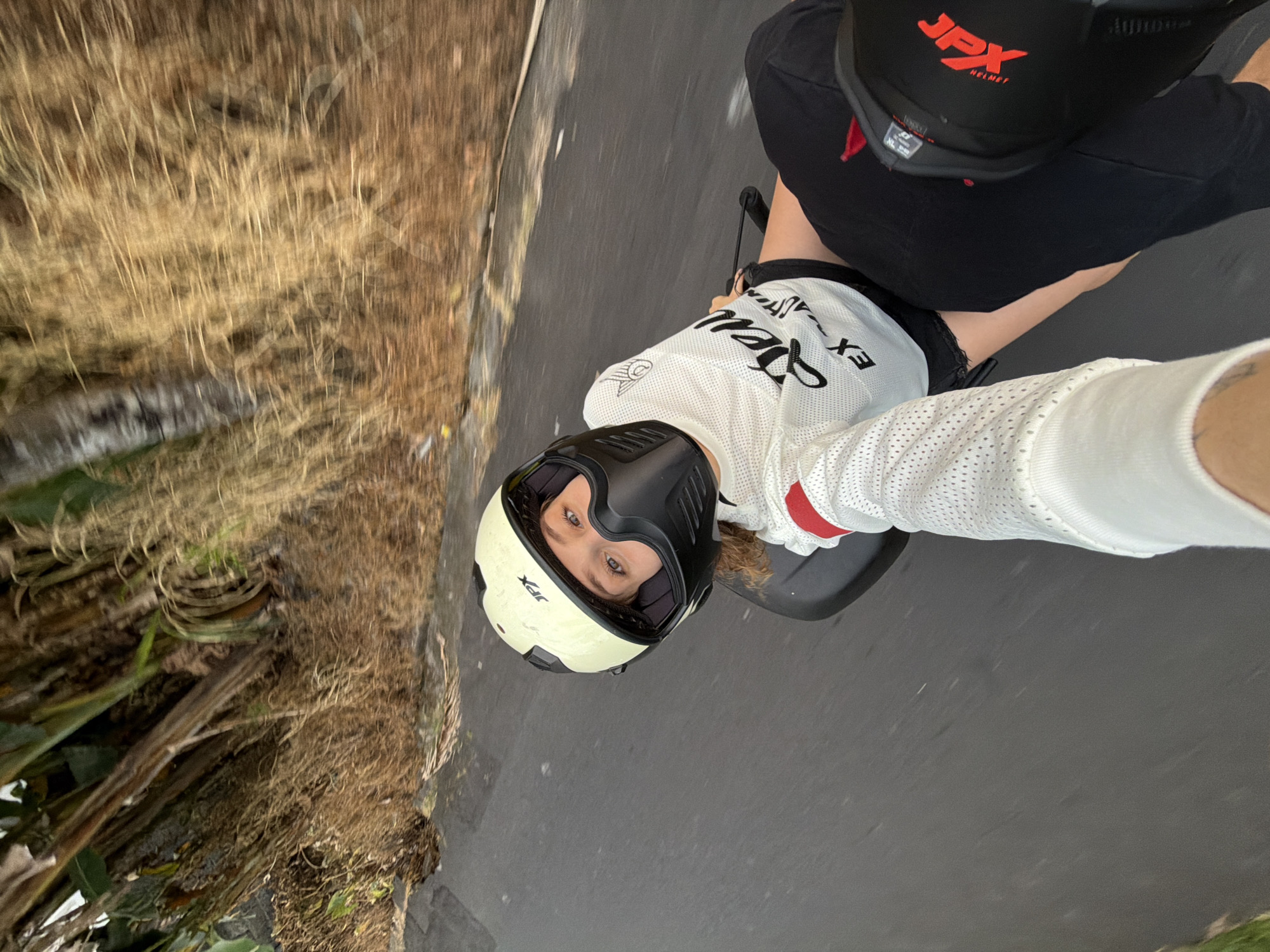 In North Kuta, Indonesia, a person is captured in a selfie video while riding what appears to be an electric scooter or motorcycle. The rider, wearing a white and yellow helmet with a black face shield, is looking directly at the camera. They are also wearing a white, mesh-like shirt with black text and red accents on the sleeves, black shorts, and black knee pads with red markings. The background shows a road with dry, tall grass and palm trees lining the right side. The motion blur indicates the scooter is in motion, moving along the paved road.