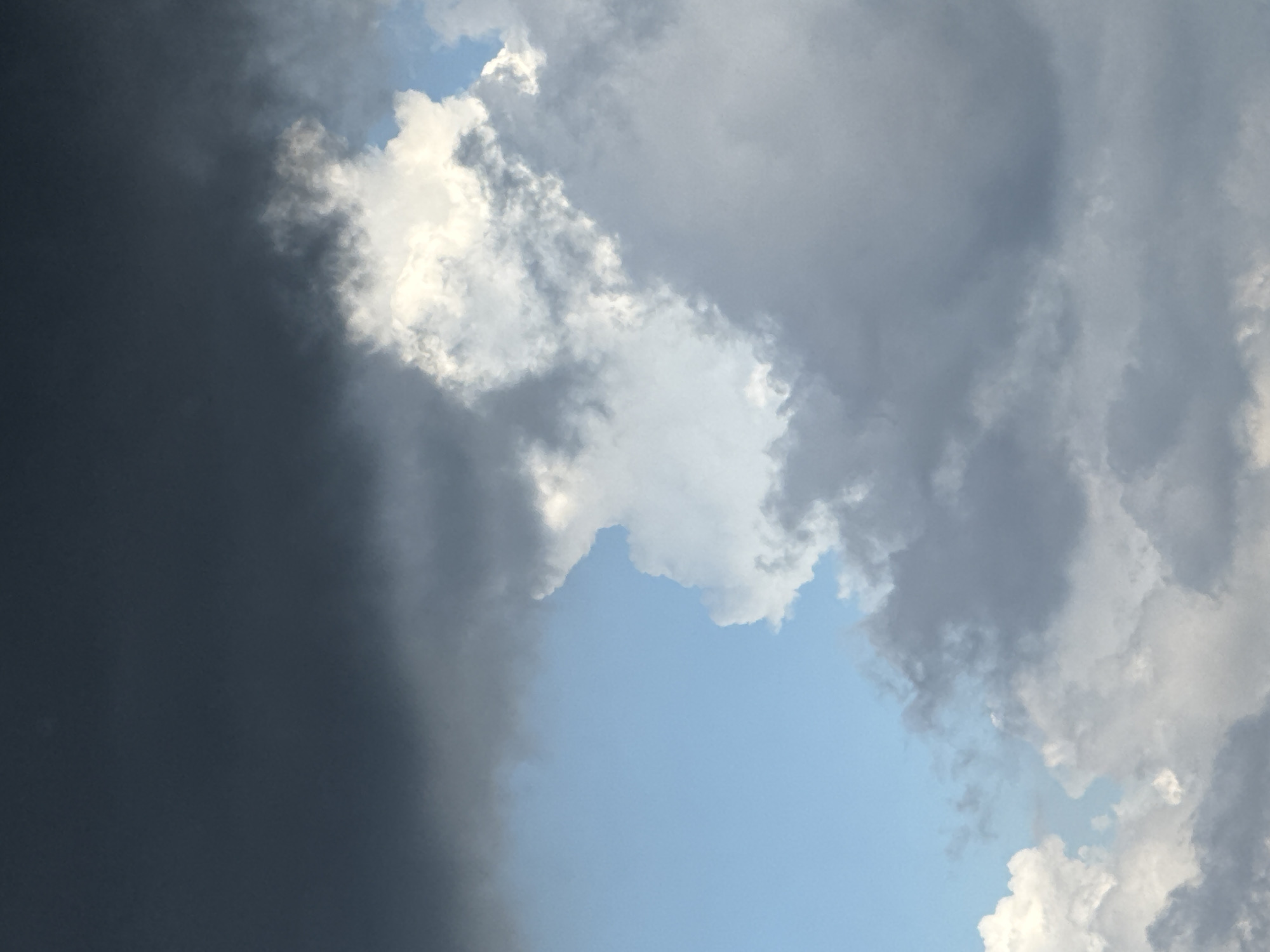 The image captures a dynamic sky with a mixture of dark storm clouds and bright white cumulus clouds. A patch of clear blue sky is visible in the upper left quadrant. The clouds are dense and textured, with varying shades of gray and white, suggesting impending rain or recent rainfall. The scene appears to be an outdoor environment, likely a landscape or cityscape given the context of Đa Lộc, Vietnam. No people or specific objects are identifiable within the frame, focusing solely on the atmospheric conditions. The lighting suggests it could be late afternoon or early evening, with the sun partially obscured by the clouds. The overall impression is one of dramatic weather.