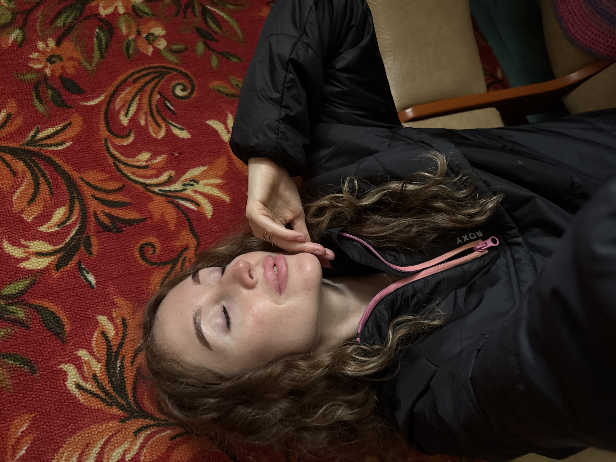 An overhead shot depicts a young woman lying down in an indoor setting, likely in Maksimovo, Russia. Her head rests on a vibrant red carpet adorned with intricate floral and paisley patterns in shades of yellow, orange, green, and black. Her long, wavy, light brown hair is spread around her head.
The woman's eyes are closed, and her lips are slightly parted, suggesting a moment of peaceful rest, sleep, or deep thought. She wears a black puffer jacket with a bright pink zipper, and the brand name "ROXY" is visible near the collar. Her right hand is gently placed near her face, with multiple rings visible on her fingers.
In the background, parts of a beige upholstered armchair or sofa with a wooden armrest are visible, indicating the presence of furniture. Glimpses of a darker green object and a woven pink and purple item can also be seen. The overall atmosphere is calm and intimate, focusing on the woman's serene expression against the richly patterned carpet. The lighting is soft and does not indicate a specific time of day.