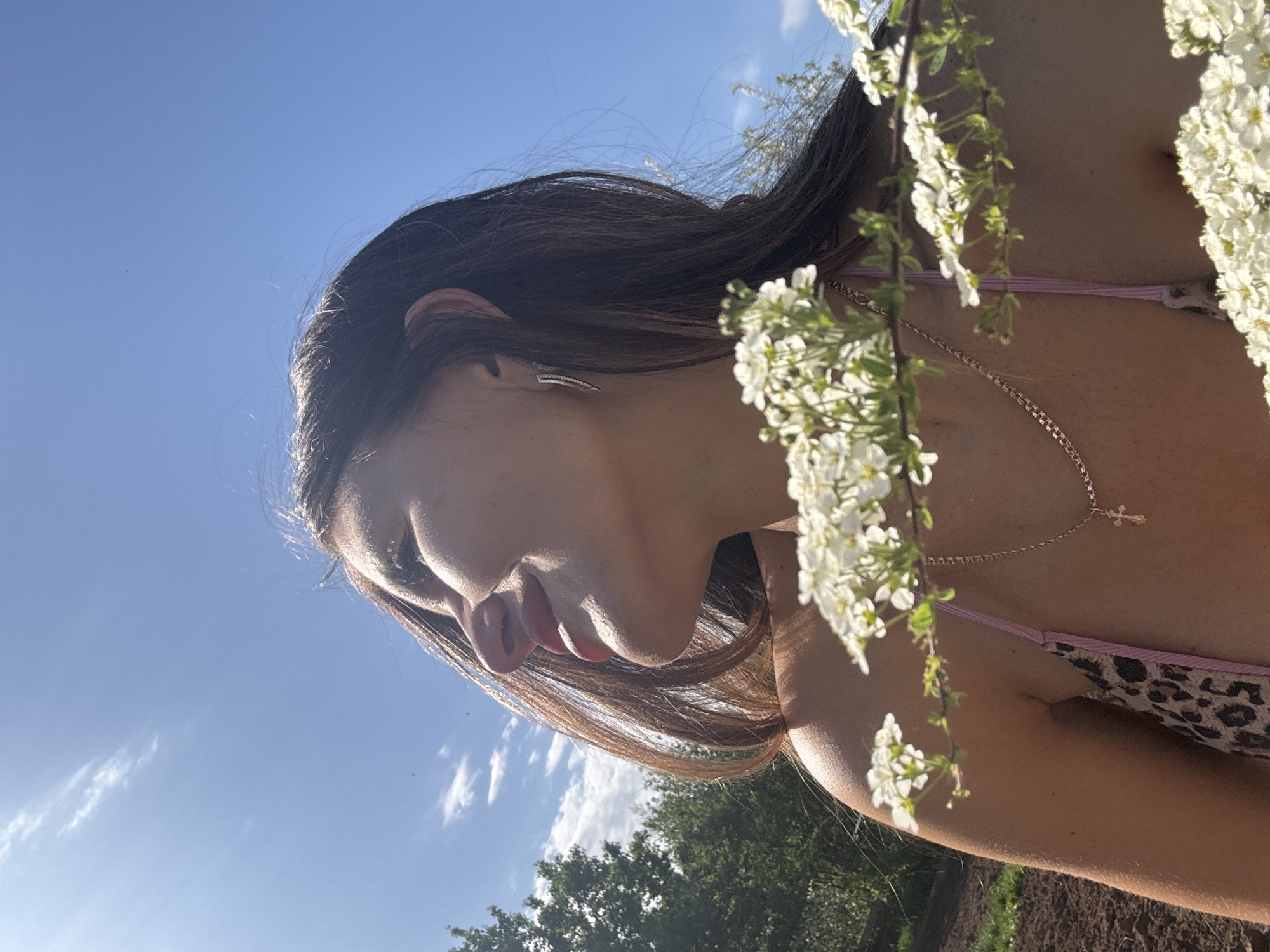 A medium shot of a woman in a bikini standing outdoors in Dubovaya, Russia, on a sunny day. The woman is facing left with her head slightly lowered, positioned at an angle so that the viewer looks up at her from below. She has long, dark hair and fair skin. She is wearing a bikini with a leopard print and pink straps and a gold necklace with a cross pendant. White flowering branches hang in the foreground, partially obscuring her. In the background, the sky is blue with some wispy clouds. Green trees and brown soil are visible at the bottom of the frame.