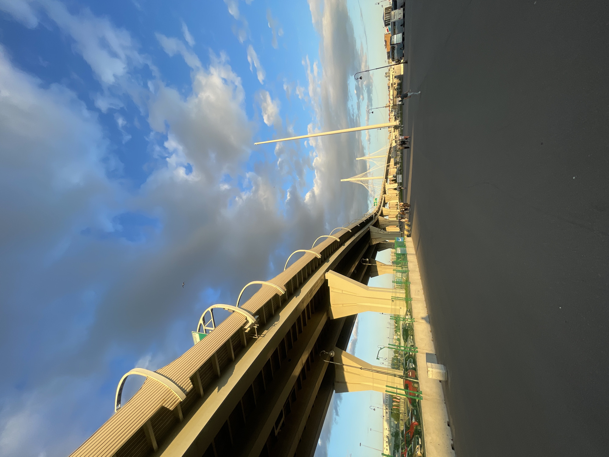 An eye-level, medium shot captures a wide asphalt road leading towards a large, modern cable-stayed bridge on a partly cloudy day. The road occupies the foreground, with a slight upward angle, while the bridge dominates the midground and background. Sunlight illuminates the scene from the left, casting warm shadows on the road and the underside of the bridge. Several concrete pillars support the elevated road structure.
On the left side of the road, a parking lot is visible, filled with various cars. A green metal fence separates the parking lot from the road. On the right side of the road, several white buses are parked. A tall, slender pole stands near the bridge, its purpose unclear.
A few people are scattered across the scene, walking on the road and sidewalks, with no discernible interactions. The sky is a vibrant blue, punctuated by dramatic white and gray clouds that create a sense of depth and movement. The overall atmosphere is calm and serene, with a hint of industrial urbanism.