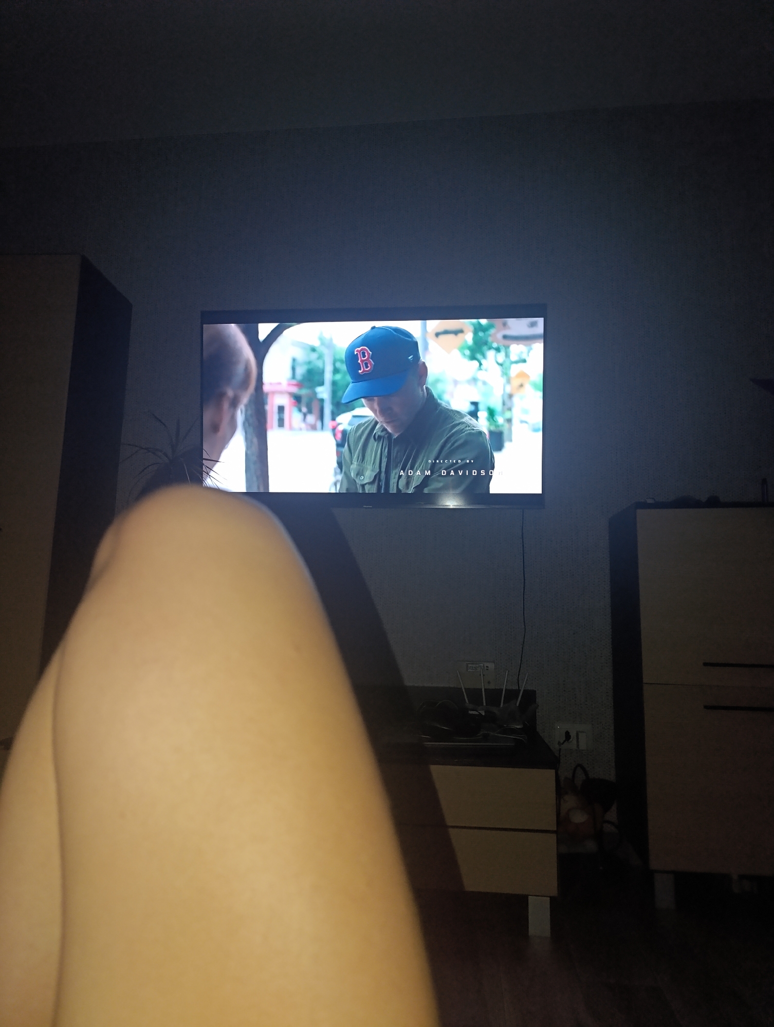 A television screen displays a scene from a movie or show. On the screen, a person wearing a blue baseball cap with a red "B" logo is visible from the chest up. The person appears to be looking downwards. Text that reads "DIRECTED BY ADAM DAVIDSON" is displayed at the bottom of the screen. In the foreground, a person's leg is visible, out of focus. The scene is set in a dimly lit room with furniture on either side of the television. A cabinet with drawers is visible to the right of the television, and a tall, dark cabinet is on the left. A plant can be seen behind the television on the left. The floor is a dark wood laminate. Electrical outlets and cords are visible on the wall behind the cabinet. The overall impression is of a domestic setting with someone watching television.