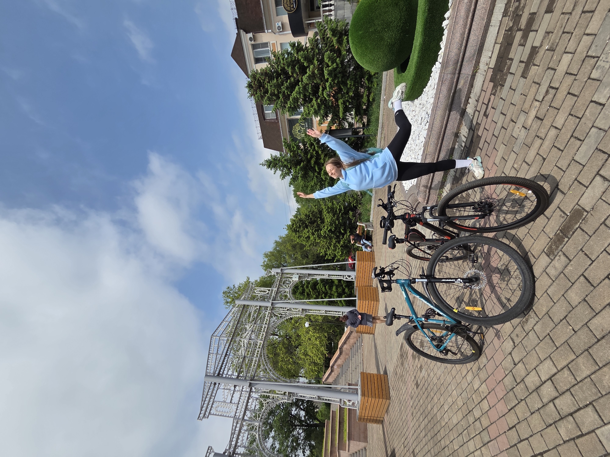 A young woman is dynamically posed atop two parked bicycles on a brick-paved area. She is wearing a light blue hoodie, black leggings, white socks, and sneakers. Her arms are outstretched to either side.
The location appears to be a public park or plaza in the city of Birobidzhan, Russia. Behind her, there is a decorative metal archway structure, some green trees, and a building with a commercial sign. A small group of people are sitting on a bench in the background. Next to her there are some white rocks and artificial bushes. The sky is blue with scattered white clouds.