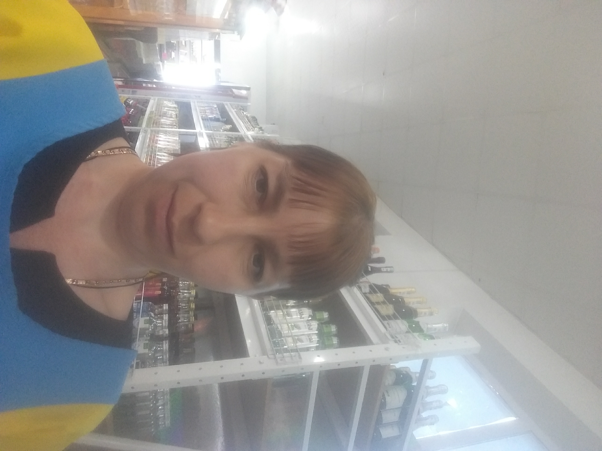 A woman is taking a selfie in what appears to be a store. She is wearing a top with blue and yellow on the shoulder and a black neckline, along with a chain necklace. Behind her are shelves stocked with various items, including what appear to be bottles and glassware. The lighting is bright, and the setting suggests a commercial establishment. The location is Бородулиха ауылдық округі, Kazakhstan.