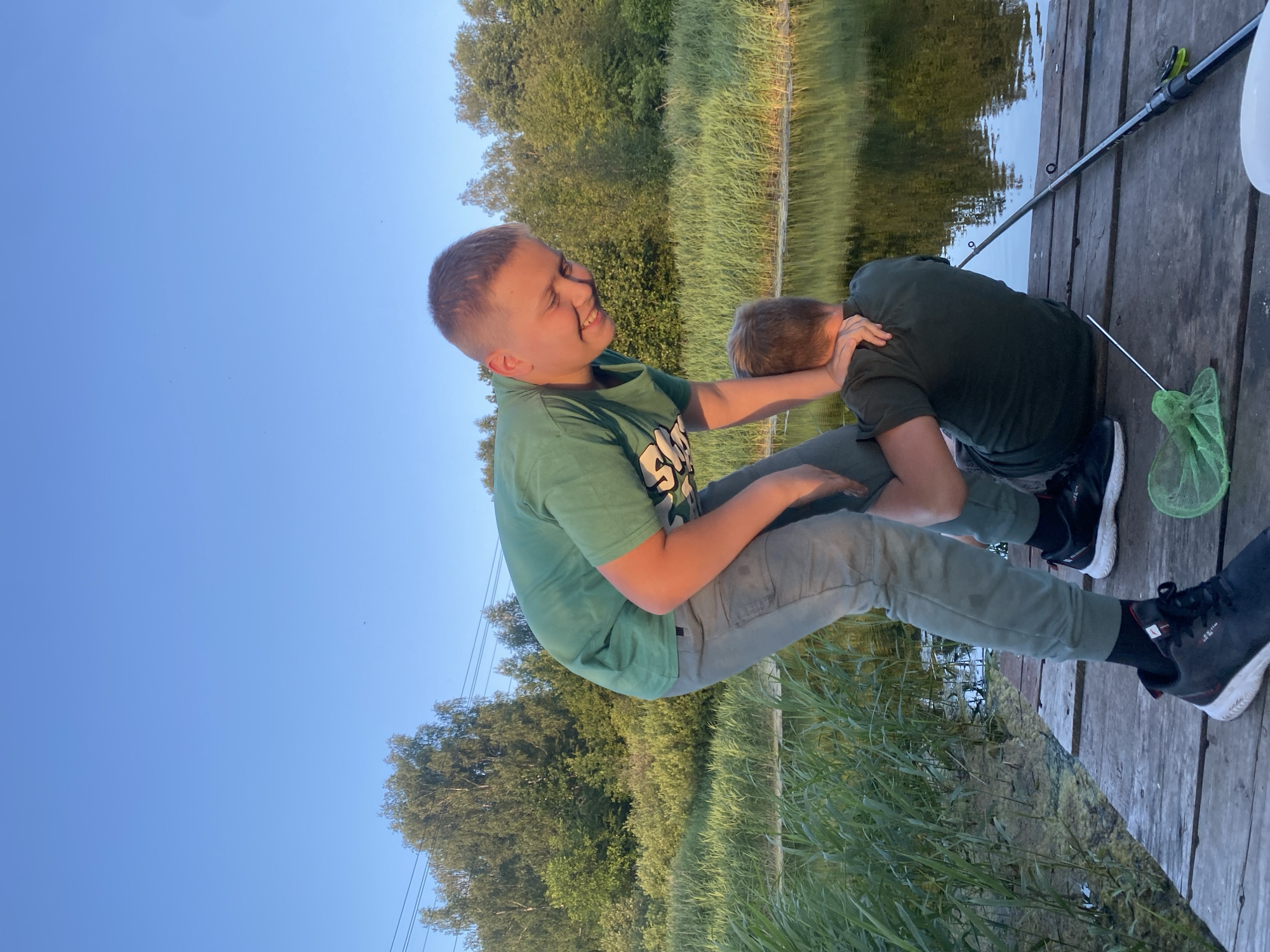 Two young boys are on a wooden pier next to a body of water. The boy on the left, wearing a green t-shirt and grey pants, is leaning over and holding the arm of the boy on the right, who is wearing a dark green t-shirt and sitting on the pier. The boy on the left is smiling. A fishing rod lies on the pier next to the sitting boy. A green fishing net is on the pier in front of the boy on the left. The background shows lush green vegetation and trees under a clear blue sky. The scene appears to be in a rural area, possibly near Afanasyevo, Russia, given the context of the location request, and the environment suggests a summer day.