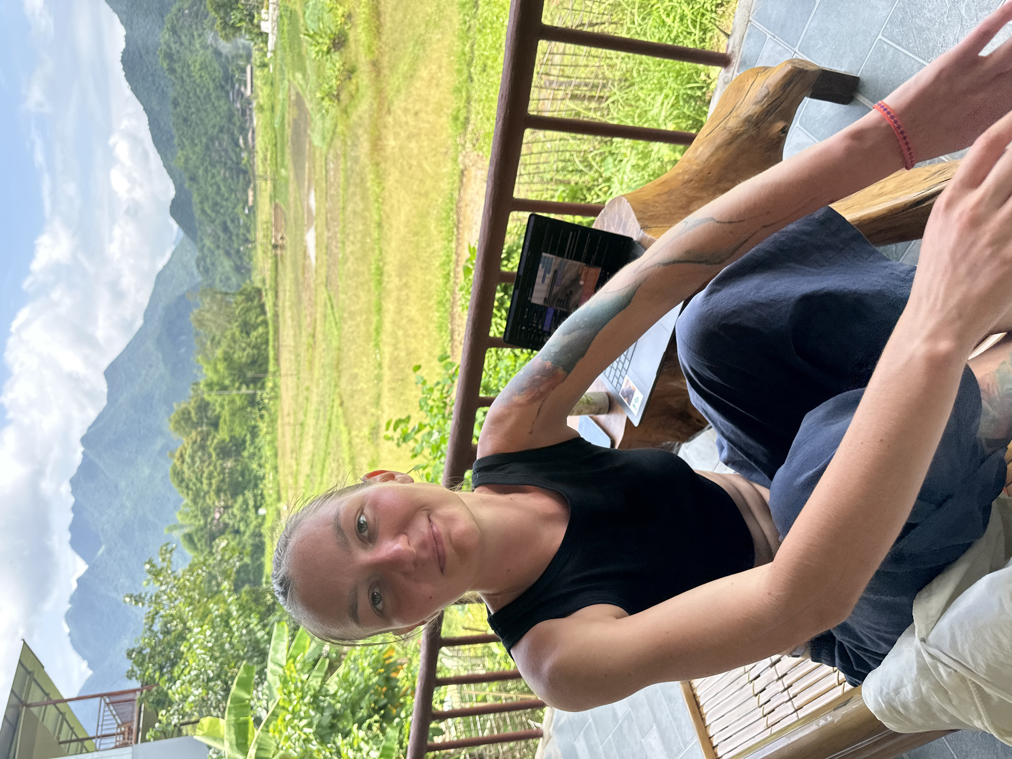 A young woman with blonde hair pulled back sits on a wooden bench on a balcony overlooking a rural landscape. She is wearing a black tank top and dark blue pants. Her left arm has a tattoo. A laptop is open on a wooden side table next to her. The background shows lush green mountains and rice paddies. The sky is cloudy. The bench is made of bamboo and has a white cushion. A railing runs along the edge of the balcony. The location appears to be Xóm Khán, Vietnam, given the mountainous terrain and rice paddies.
