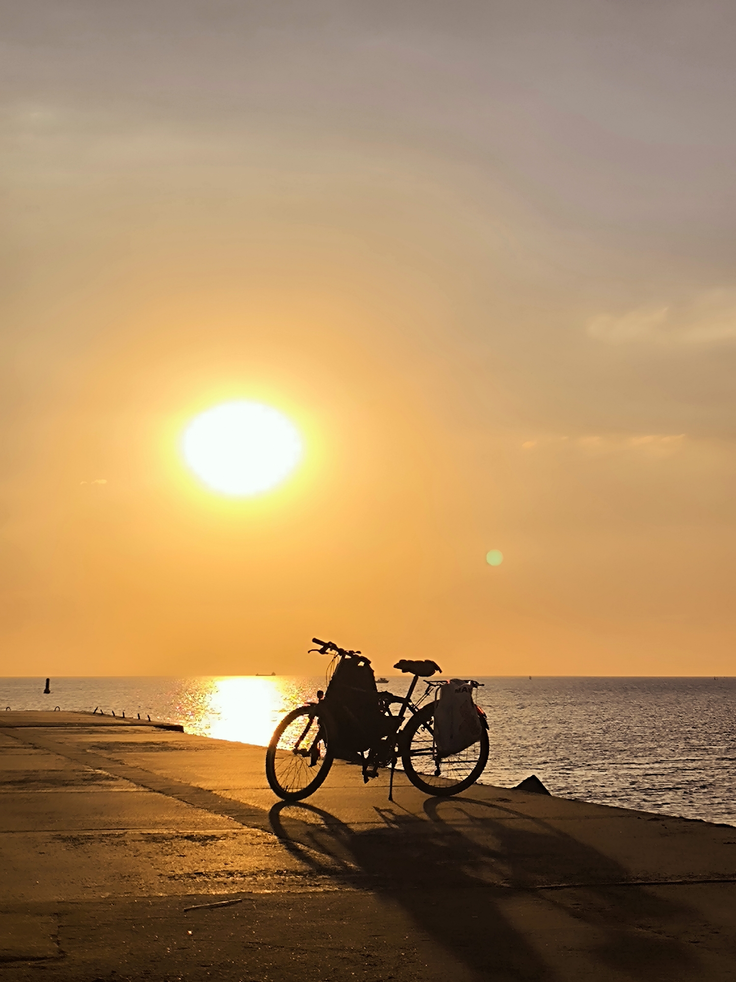 The image depicts a silhouette of a bicycle parked on a paved pier or walkway along the coast of the Baltic Sea. The sun is setting, casting a warm, golden-orange glow across the sky and reflecting on the water. The bicycle has two bags attached, one on the front handlebars and another on the rear rack. In the distance, a few ships are visible on the horizon. The scene appears serene and peaceful, with the natural beauty of the sunset dominating the composition. Given the context of Riga, Latvia, this location could be along the Daugava River estuary or the coast of Jurmala, a popular resort city near Riga.