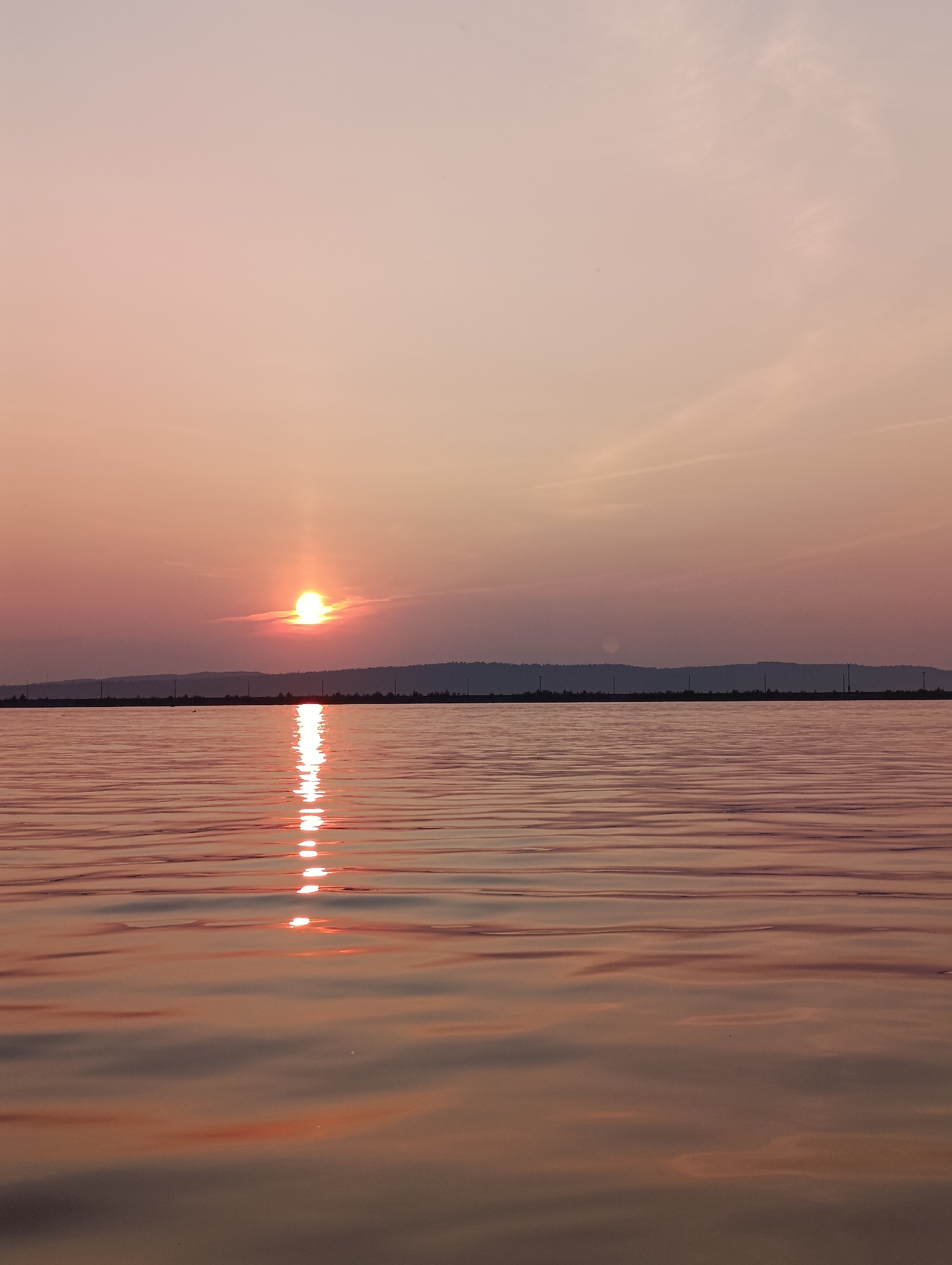 This image captures a serene sunset over the Kama River in Chaikovsky, Russia. The sun, a bright orange orb, is partially obscured by a thin layer of clouds, casting a warm, pinkish glow across the sky. A reflection of the sun shimmers on the rippling surface of the river, creating a path of light that stretches towards the viewer. The horizon is marked by a dark, silhouetted line of trees and hills, with several utility poles scattered along the distant shoreline. The water itself exhibits gentle waves, reflecting the soft hues of the sky and the sun. The overall impression is one of tranquility and natural beauty.
