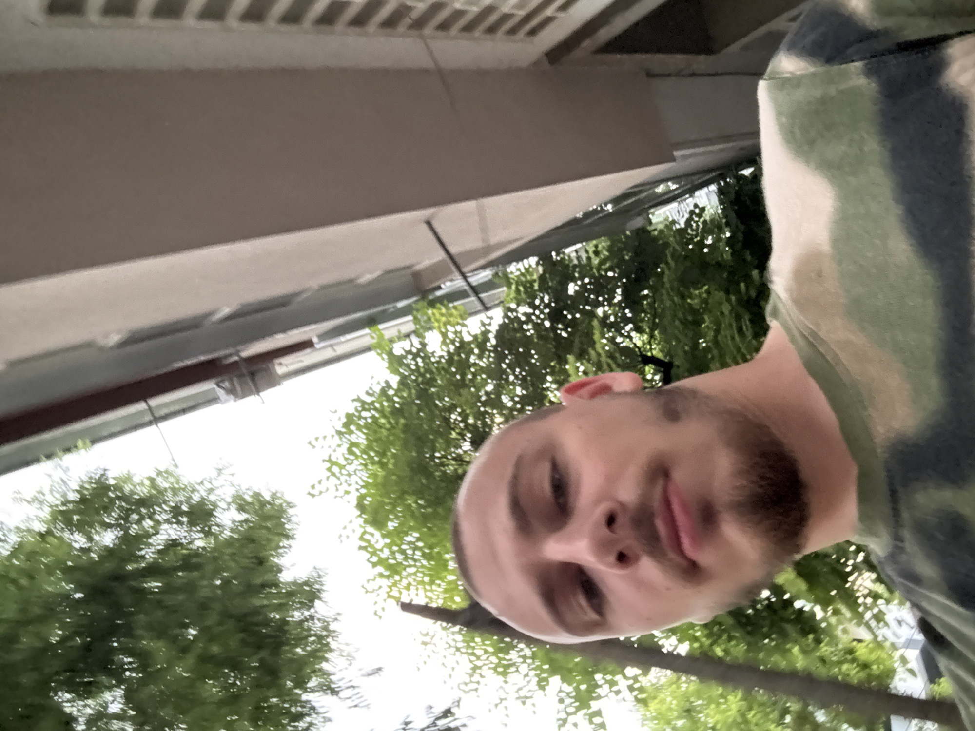 The image is a low-angle, close-up outdoor selfie of a man, likely taken looking upwards. The man, who appears to be an adult, has a shaved head or very short hair and a neatly trimmed beard. He is wearing a t-shirt with a green and white camouflage or tie-dye pattern. His expression is neutral or slightly pensive, with his eyes looking towards the camera.
In the background, lush green trees with abundant foliage are visible, suggesting a natural or park-like setting. Above the trees, a bright, clear sky indicates daytime. To the left and upper part of the frame, a concrete or stone building structure, possibly an overhang, balcony, or part of a ceiling, is visible. Thin dark lines, possibly wires or supports, extend from this structure.
The scene depicts a bright, clear day, and the setting appears to be an urban or suburban outdoor environment, specifically in Stara Zagora, Bulgaria. There is no visible text in the image.
