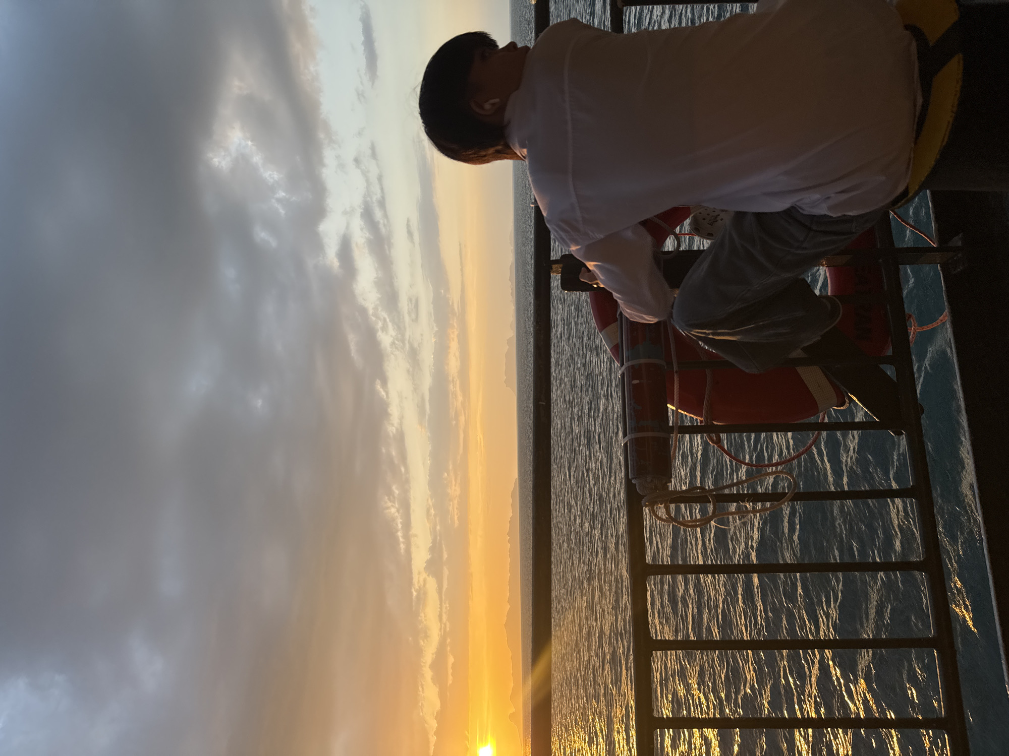 A person, silhouetted against a vibrant sunset over the ocean, sits on a yellow cushion on the deck of a ferry. The person, with short dark hair and wearing a white button-down shirt and jeans, faces away from the camera, looking out at the horizon. The sun, a bright orb of orange, casts a golden sheen on the rippling water and illuminates a distant island chain. A black metal railing runs along the foreground, separating the deck from the water. A red life preserver with white stripes is attached to the railing, with a rope coiled around it. The sky is a mix of soft orange and gray clouds. The scene appears to be on a ferry traveling between islands in Thailand, given the tropical island silhouettes and the presence of a life preserver common on ferries.