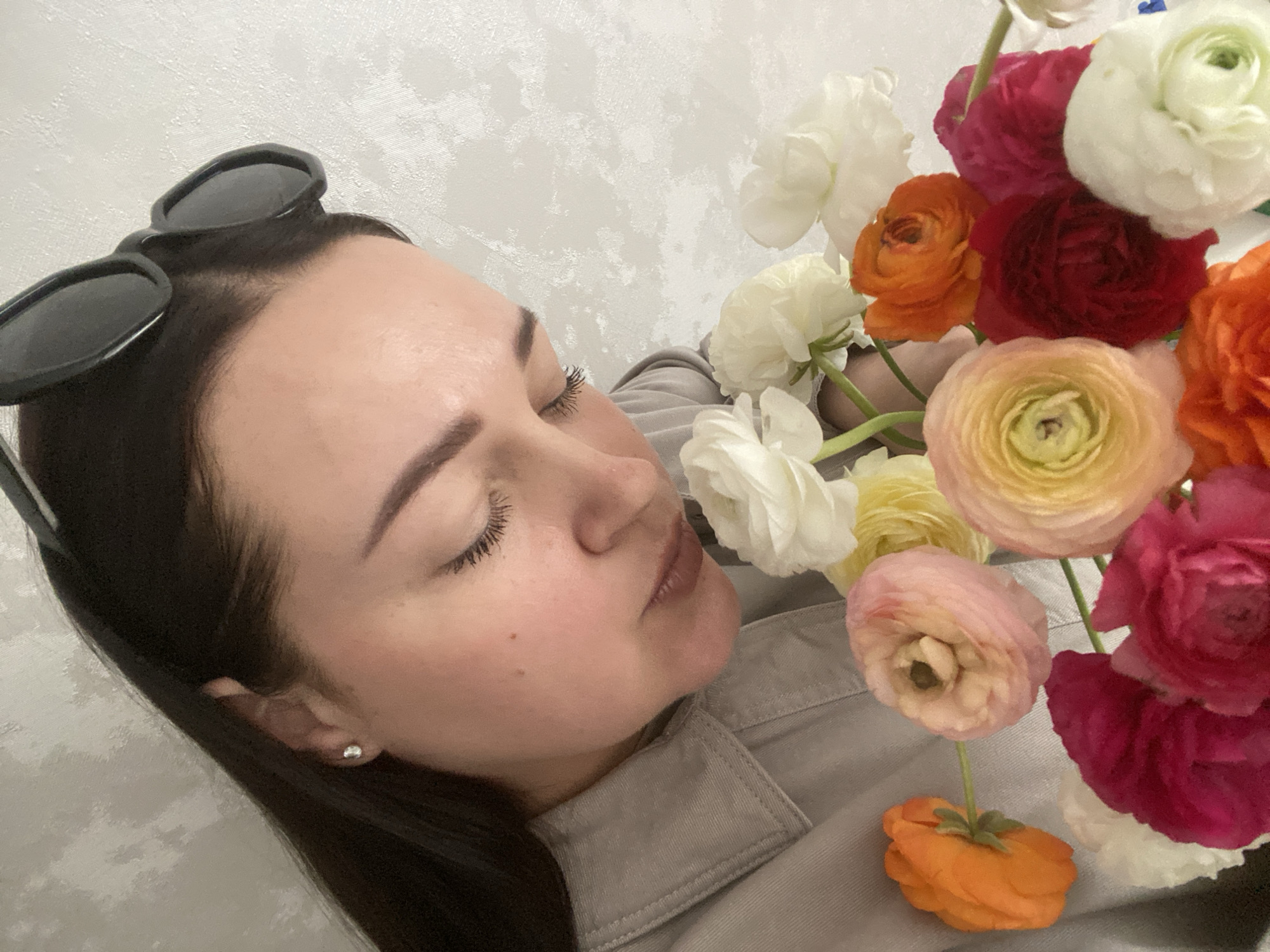 A close-up, angled portrait captures a woman with her eyes closed, appearing to rest her face near a vibrant bouquet of flowers. The scene evokes a sense of peace and appreciation.
The woman, with fair skin and dark hair, is positioned with her head tilted, revealing the right side of her face in profile. Her eyes are gently closed, and long, dark eyelashes are visible. Her lips are slightly parted, and she wears a small, silver stud earring on her visible earlobe. Black sunglasses are perched on top of her head, resting in her dark hair. She is wearing a light grey or beige collared shirt or jacket, the fabric of which is visible around her neck and shoulder.
A prominent bouquet of ranunculus flowers fills the right side of the frame. The flowers display a rich palette of colors, including white, bright orange, deep red, fuchsia pink, and soft yellow/peach. Some individual flower heads are clearly distinguishable, showcasing their characteristic ruffled petals. The flowers are arranged closely together, creating a dense and colorful cluster.
The background consists of a plain, light-colored wall with a subtle textured pattern. There is no visible text in the image. The image is set in Sochi, Russia, although no specific location cues are visible within the frame itself.