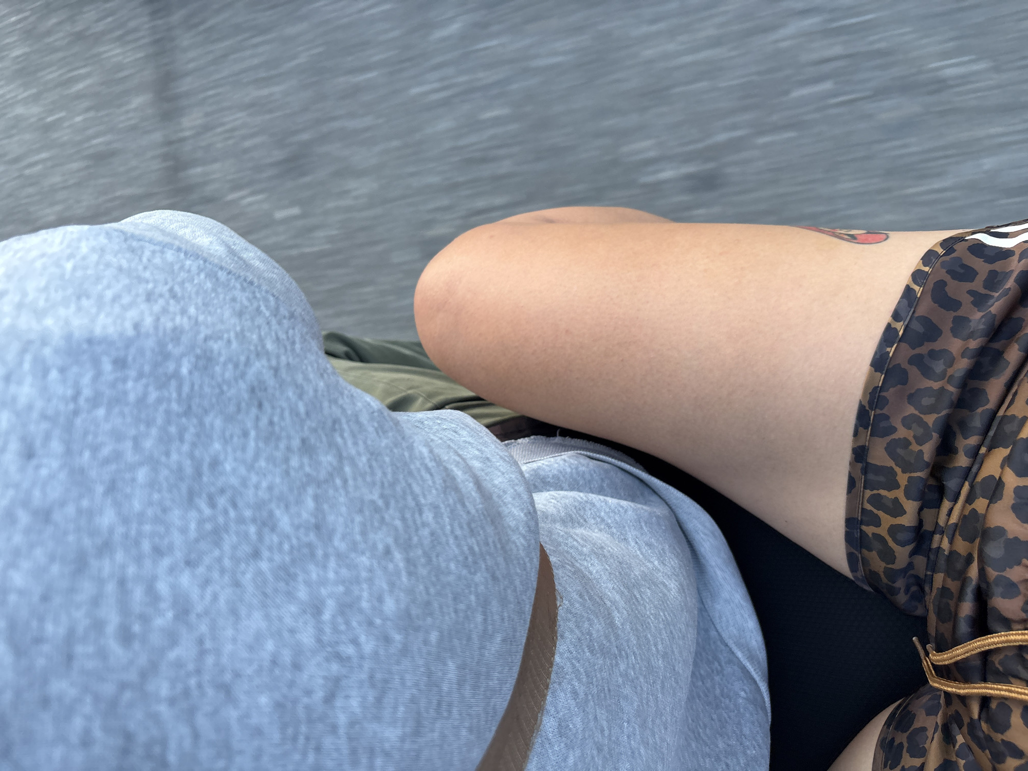 The image shows a close-up, overhead view of two individuals sitting side-by-side. The person on the left is wearing light blue sweatpants or joggers, with the fabric appearing soft and textured. Their leg is bent, and the material folds around their knee. To the right, the second person is wearing shorts with a leopard print pattern in shades of brown and black. A glimpse of their bare leg is visible, and there is a small tattoo on their upper thigh of a red and pink candy. The background is a blurred, greyish-blue textured surface, possibly a road or a seating area. The lighting suggests it is daytime, and the overall impression is casual and relaxed. The scene is set in Nong Pa Khrang, Thailand. No visible text is present in the image.