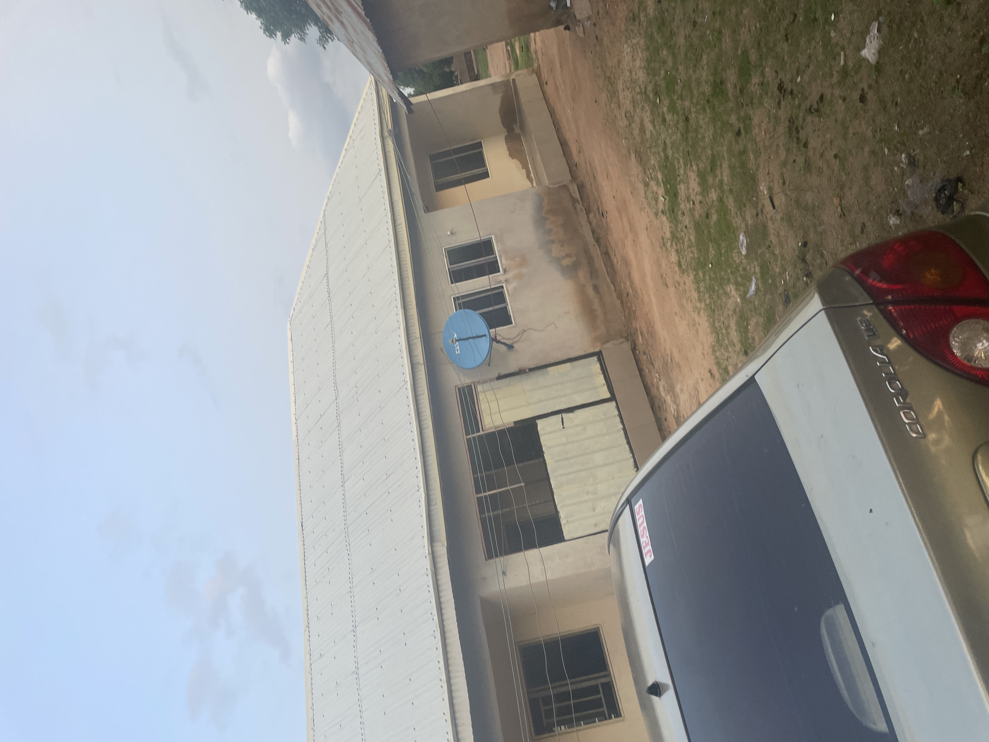 The image depicts a residential scene in Jalingo, Nigeria. In the foreground, the rear of a light-colored sedan is visible, with the word "JESUS" emblazoned on a red sticker on its rear window. The car's taillights and the "COROLLA" badging are also discernible. Behind the car, a single-story building with a corrugated metal roof dominates the midground. The building has several windows with bars and a light-colored, paneled entrance. A blue satellite dish is mounted on the exterior wall of the building. The ground surrounding the building and car is composed of reddish-brown earth and patches of dry grass, with scattered debris. Another structure, partially visible on the right side of the image, appears to have a corrugated metal roof as well. The sky above is overcast with a light blue hue and some faint clouds. The overall impression is a modest, rural or semi-urban residential setting.