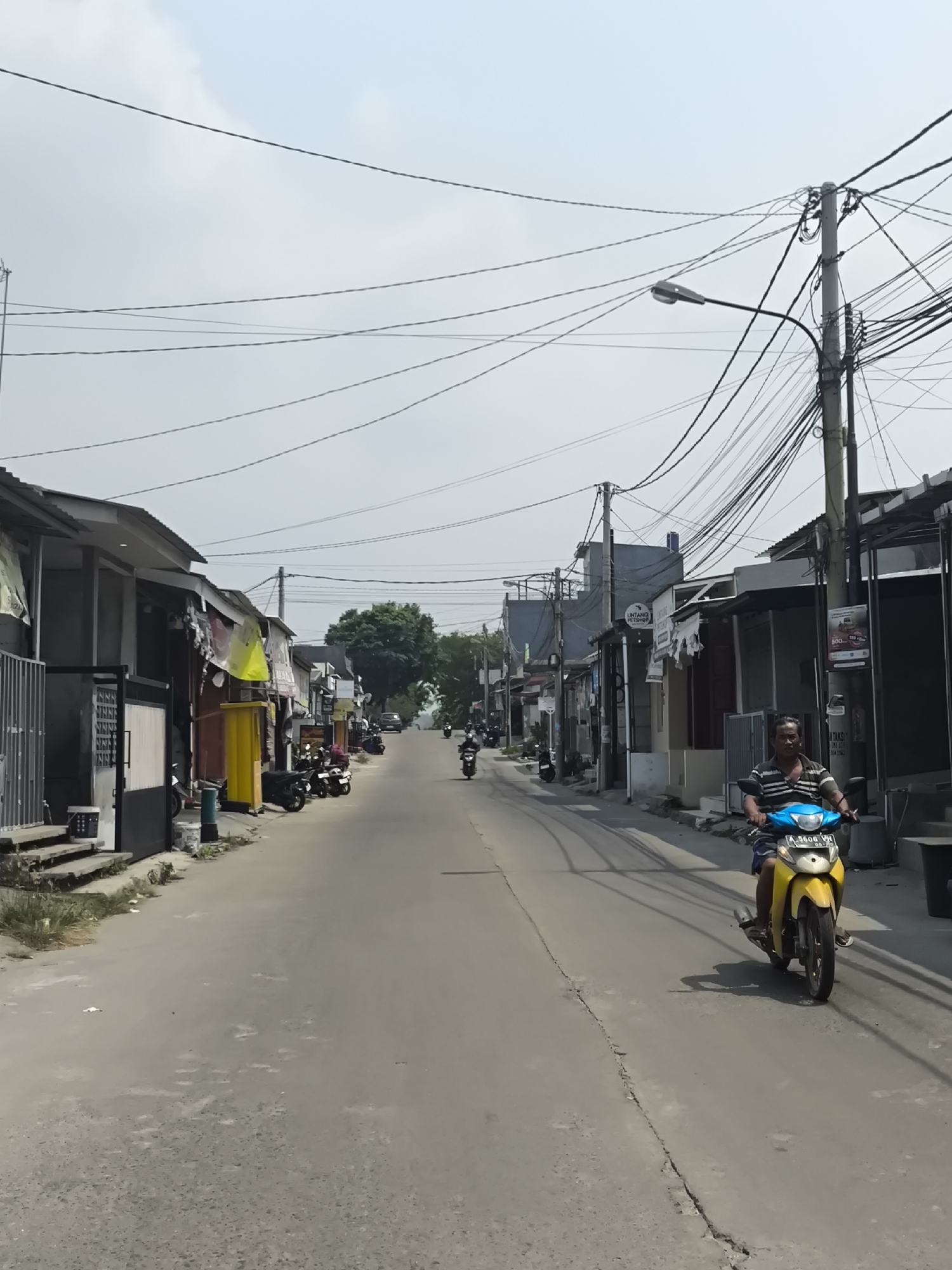 A street scene in Cilegon, Indonesia, features a person riding a yellow scooter down a paved road. The scooter has a license plate that reads "A 3606 VH." The road is lined with buildings on both sides, some of which appear to be shops or small businesses. Various motorcycles are parked along the side of the road. Overhead, a dense network of electrical wires crisscrosses the sky, supported by utility poles. The sky is overcast.