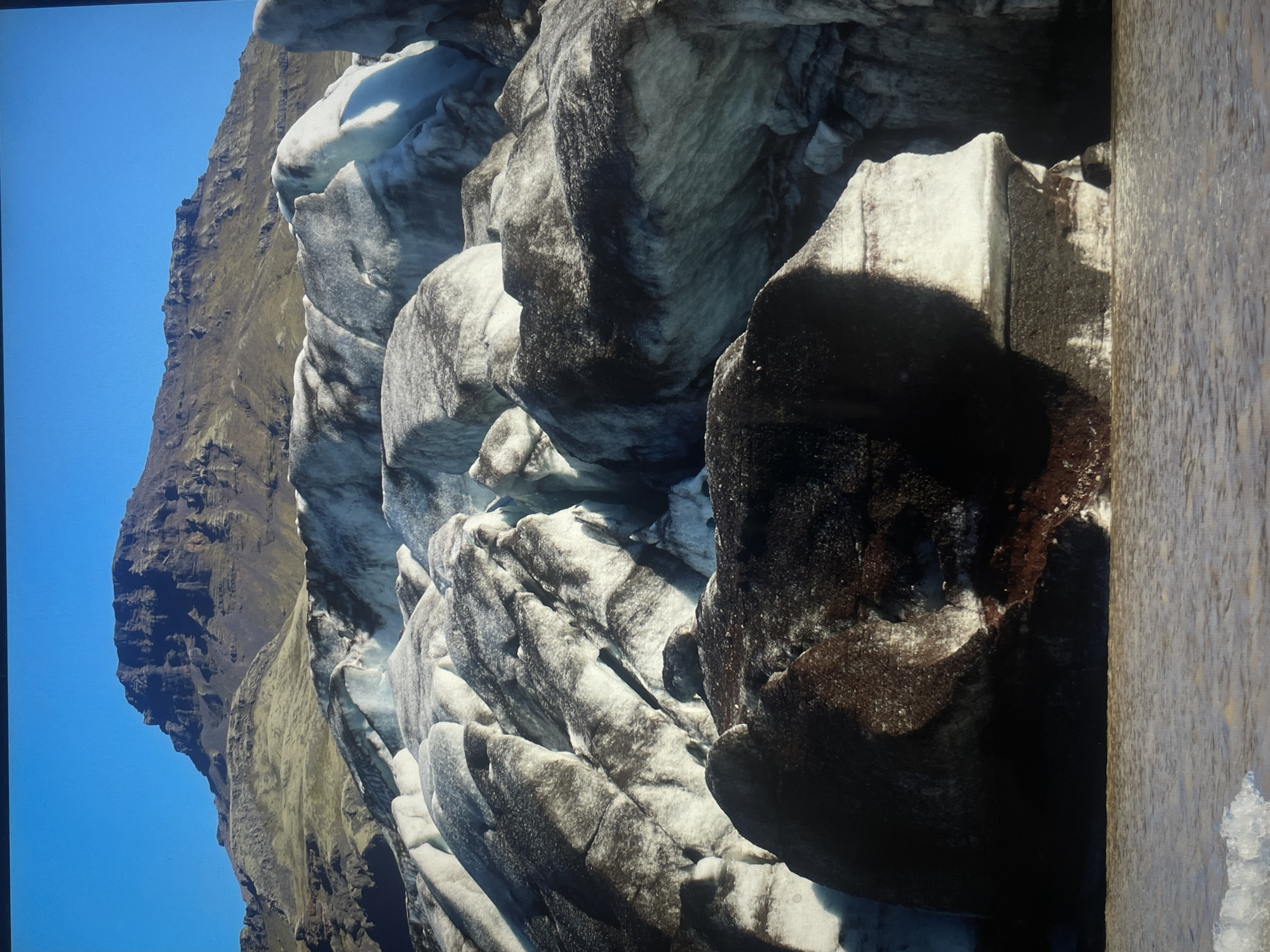 The image shows a glacier landscape with a mountain range in the background under a clear blue sky. The glacier is composed of large formations of ice, colored white and dark grey/brown, possibly due to sediment or volcanic ash. The mountain range appears rugged and barren. The location is unknown.