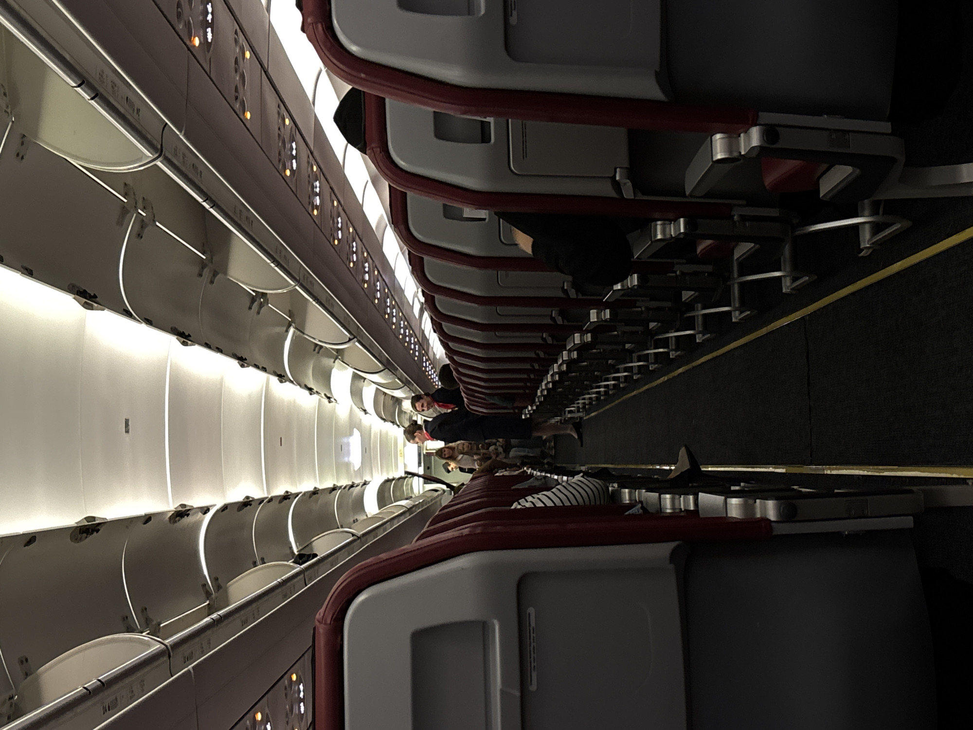 The image shows the interior of an airplane cabin. The aisle is carpeted in dark gray with a yellow line marking the edge. Rows of gray seats with red headrests and armrests line both sides of the aisle. The ceiling of the cabin is white with integrated lighting. In the distance, two women in flight attendant uniforms are standing in the aisle. One is wearing a dark blue uniform with a red scarf, and the other is wearing a similar uniform with a red scarf and a red blazer. Several passengers are visible in the seats, some with their heads resting on the headrests. The overall impression is of a commercial aircraft cabin during boarding or pre-flight. The location is stated to be Bolshoye Savino, Russia.