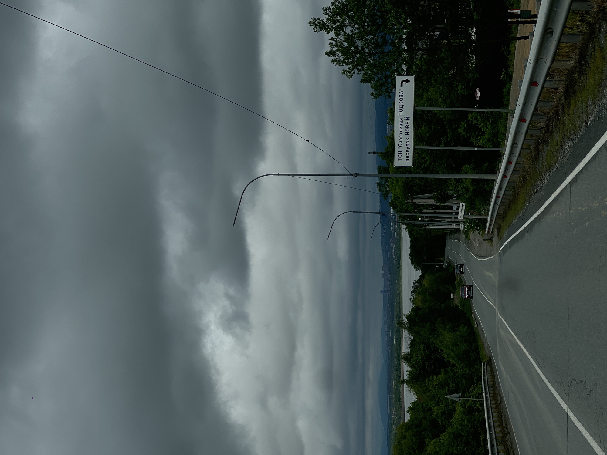 The image shows a road in Shmidtovka, Russia. A sign reads "TSN 'Happy Horseshoe' lane NEW". The road curves ahead with a barrier on the right side. Two cars are visible on the road. A line of street lights extends along the road. On the left, trees line the road with a cloudy sky above. In the distance, there is a view of a city or settlement along a body of water.