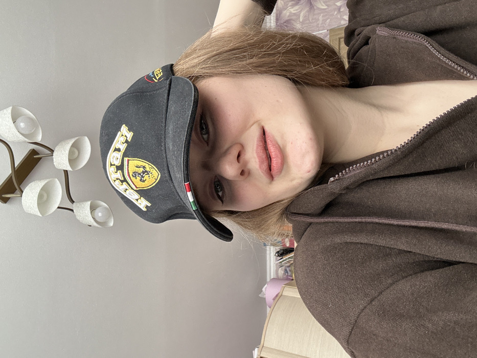 A young adult female is depicted in a residential indoor setting in Nizhnekamsk, Russia. She is shown from a low-angle perspective, facing the camera with a neutral expression and a slight smile. Her hair is light brown, and she wears a black baseball cap with "Ferrari" embroidered in yellow script and the Ferrari prancing horse logo. The cap's brim displays a green, white, and red stripe pattern. She is dressed in a dark brown hooded zipper sweatshirt, partially unzipped. Her left hand is positioned behind her head, lightly touching her hair. The background features a white ceiling with a five-arm ceiling chandelier, equipped with exposed bulbs and textured white shades. To the left, a light-colored wooden furniture piece, a pink container, and various items are partially visible. To the right, a patterned wall covering in a light purple hue is discernible.