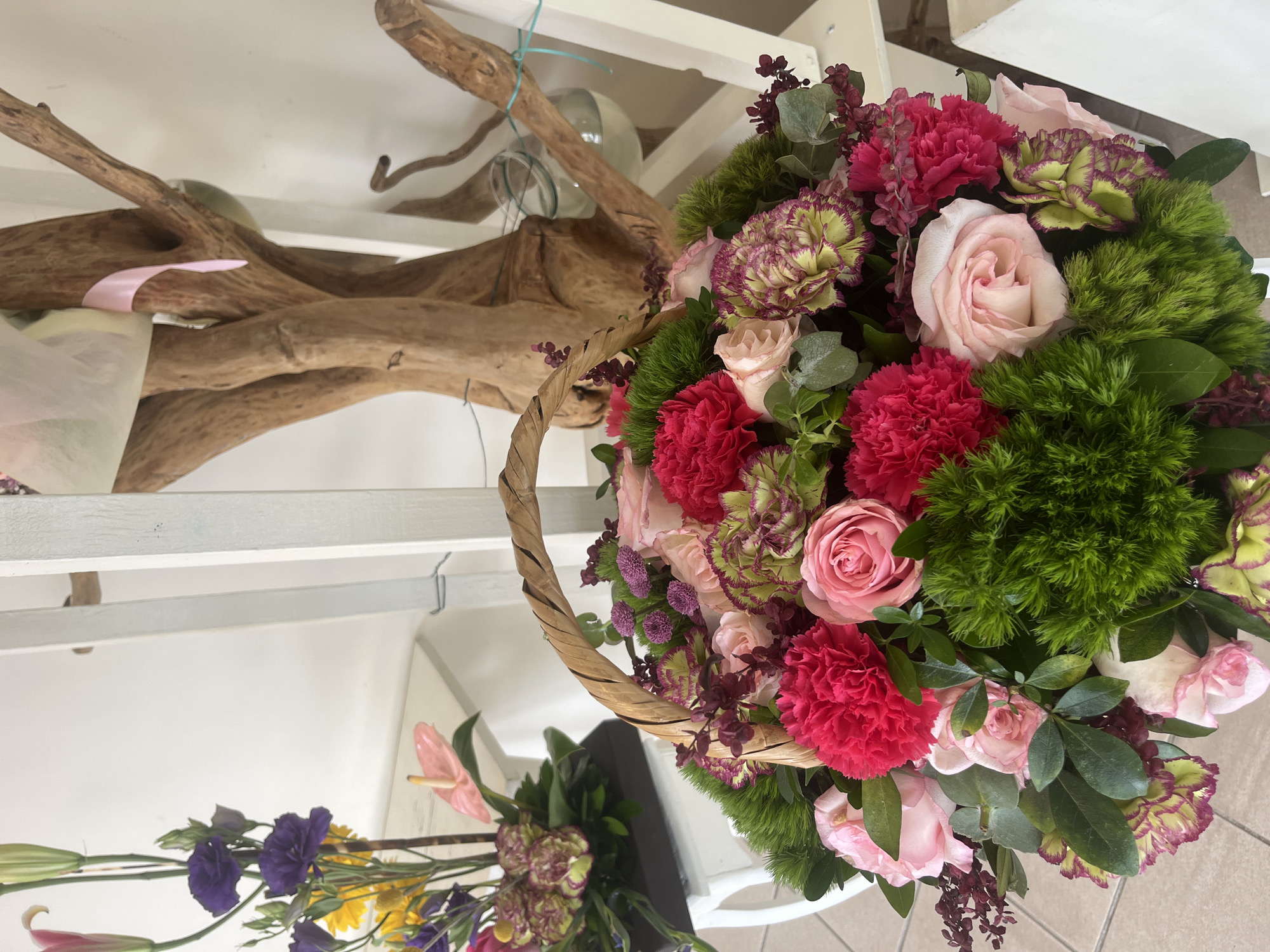The scene is an indoor floral arrangement, likely within a shop or studio in Puebla, Mexico. The primary focus is a basket overflowing with flowers, including roses, carnations, and green filler. A dried branch structure sits behind the basket, possibly as part of the shop's decor. Additional flowers, including lilies and other blooms, are partially visible below. The setting suggests a florist or a shop specializing in floral arrangements.